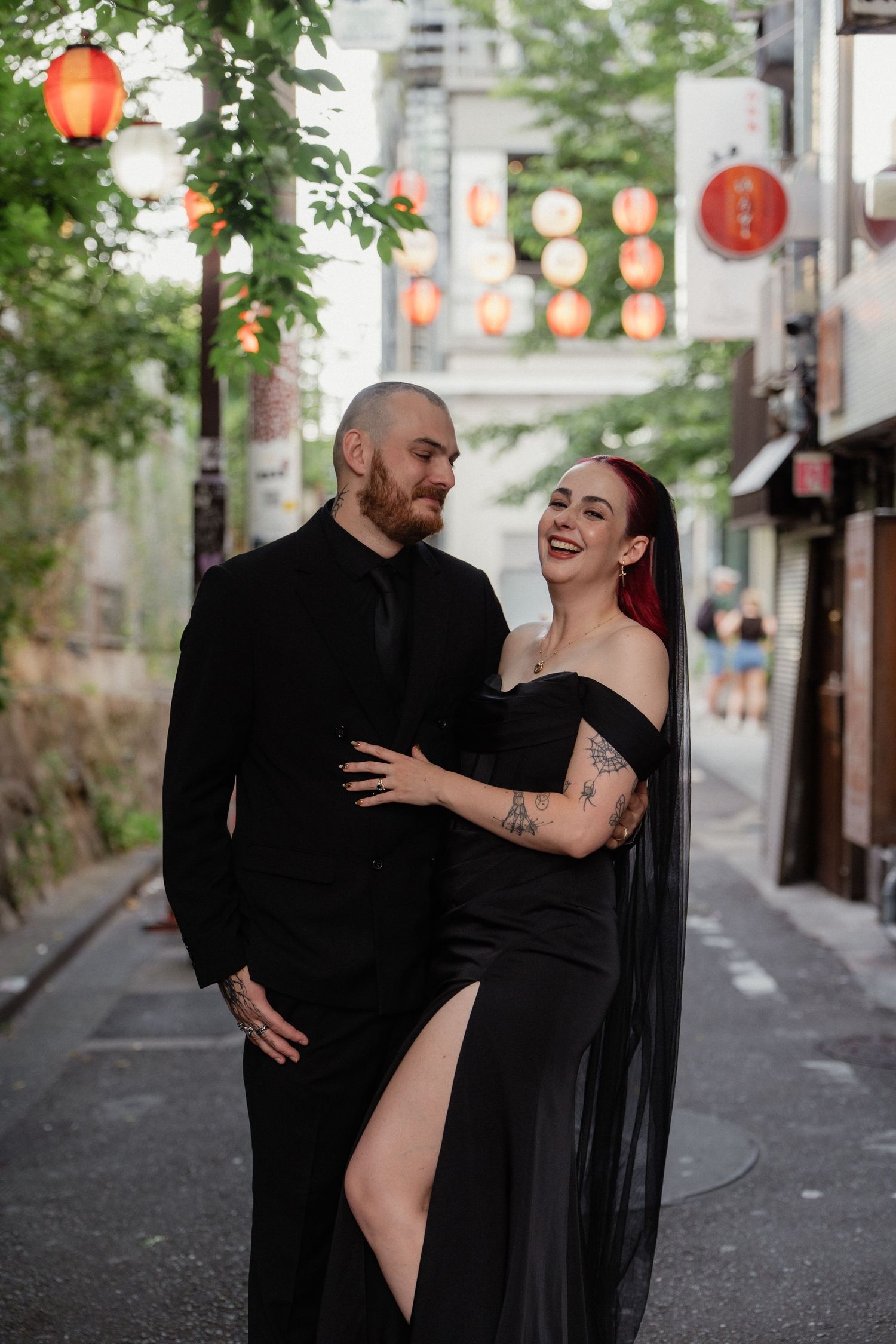 Couple in matching black formal attire embracing on a Japanese street lined with red lanterns.