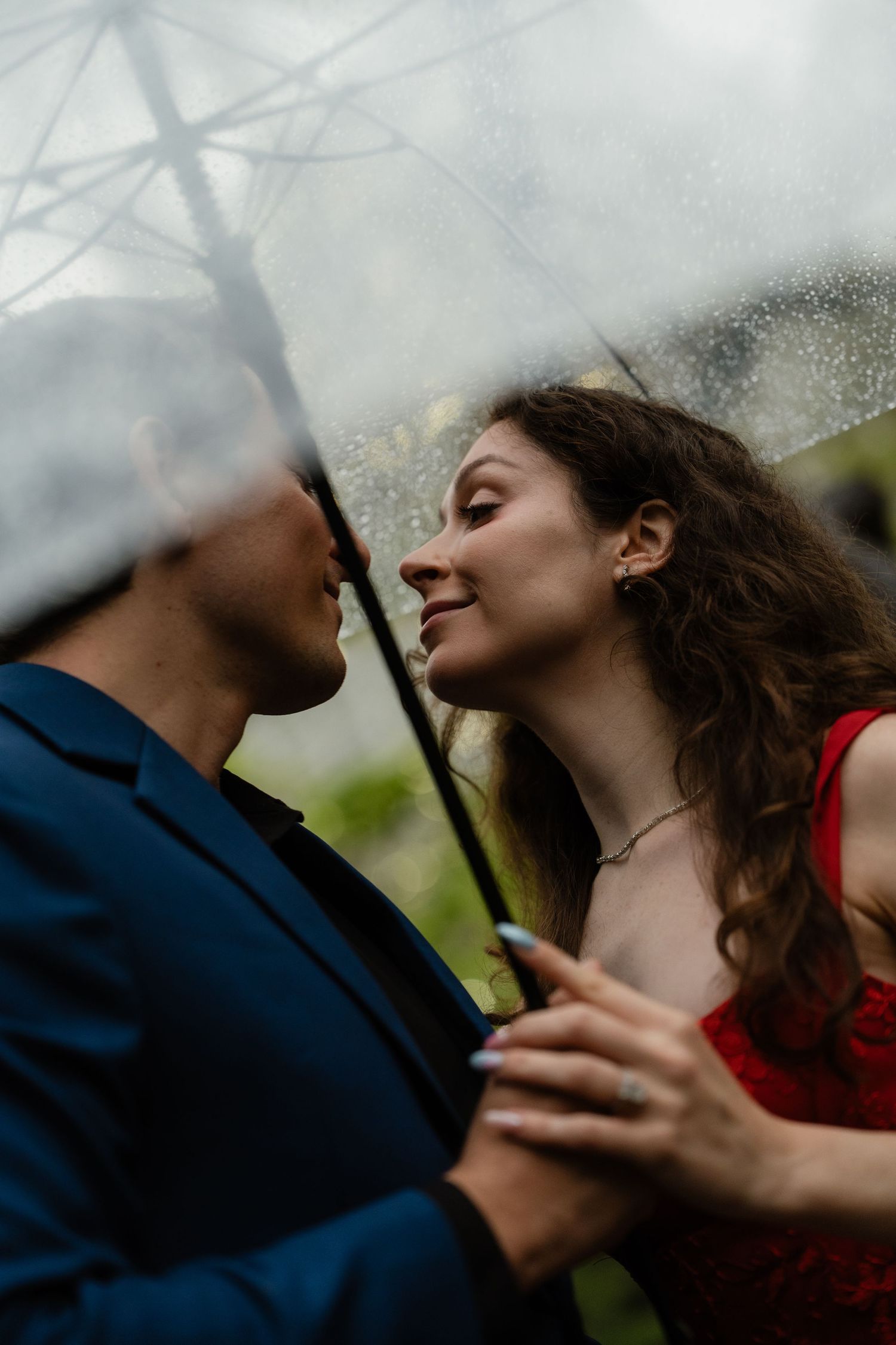 Romantic couple sharing an intimate moment under a clear umbrella in the rain, woman in red dress smiling.