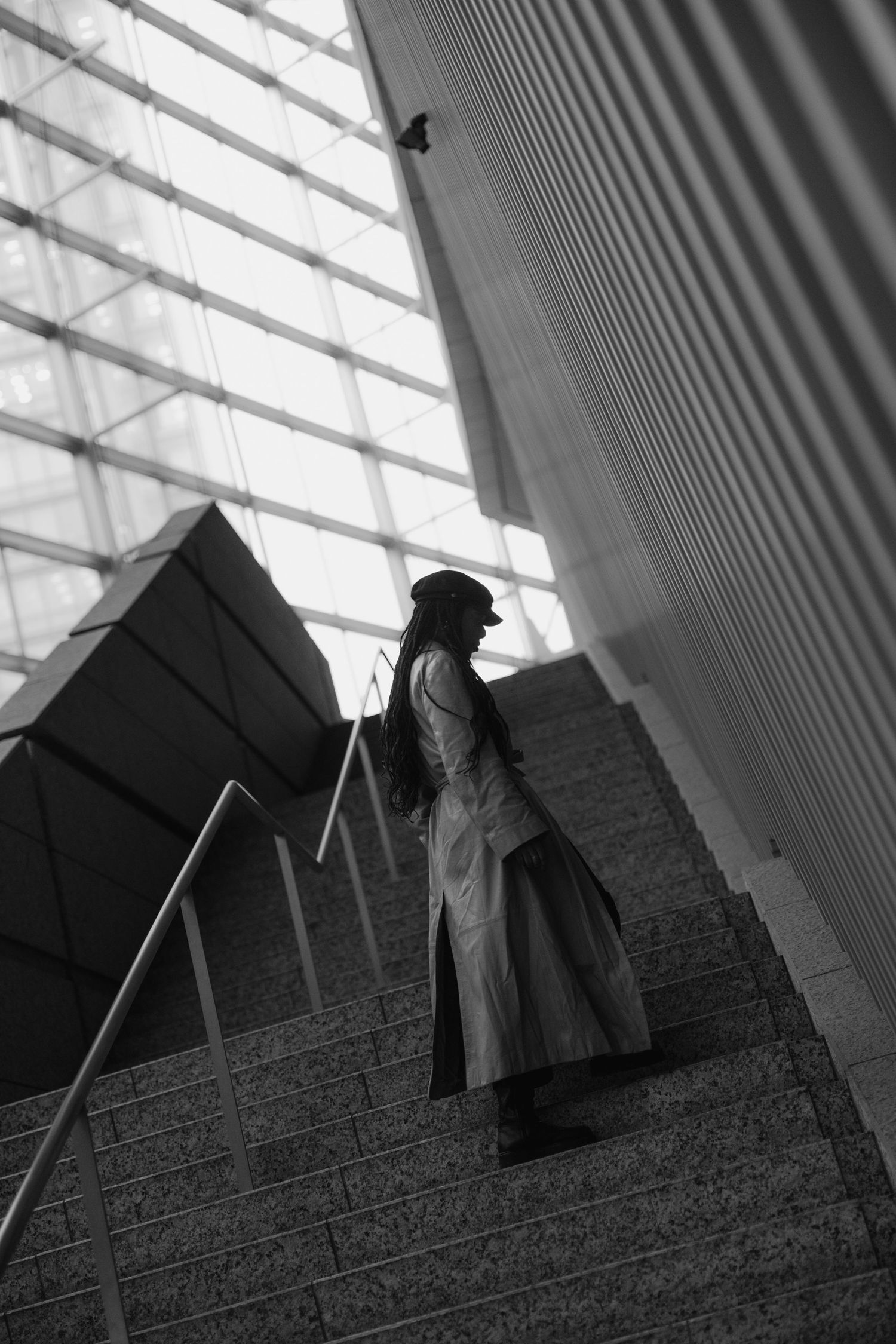 Black and white photo of a woman in a long coat ascending stairs in a modern building with large windows.
