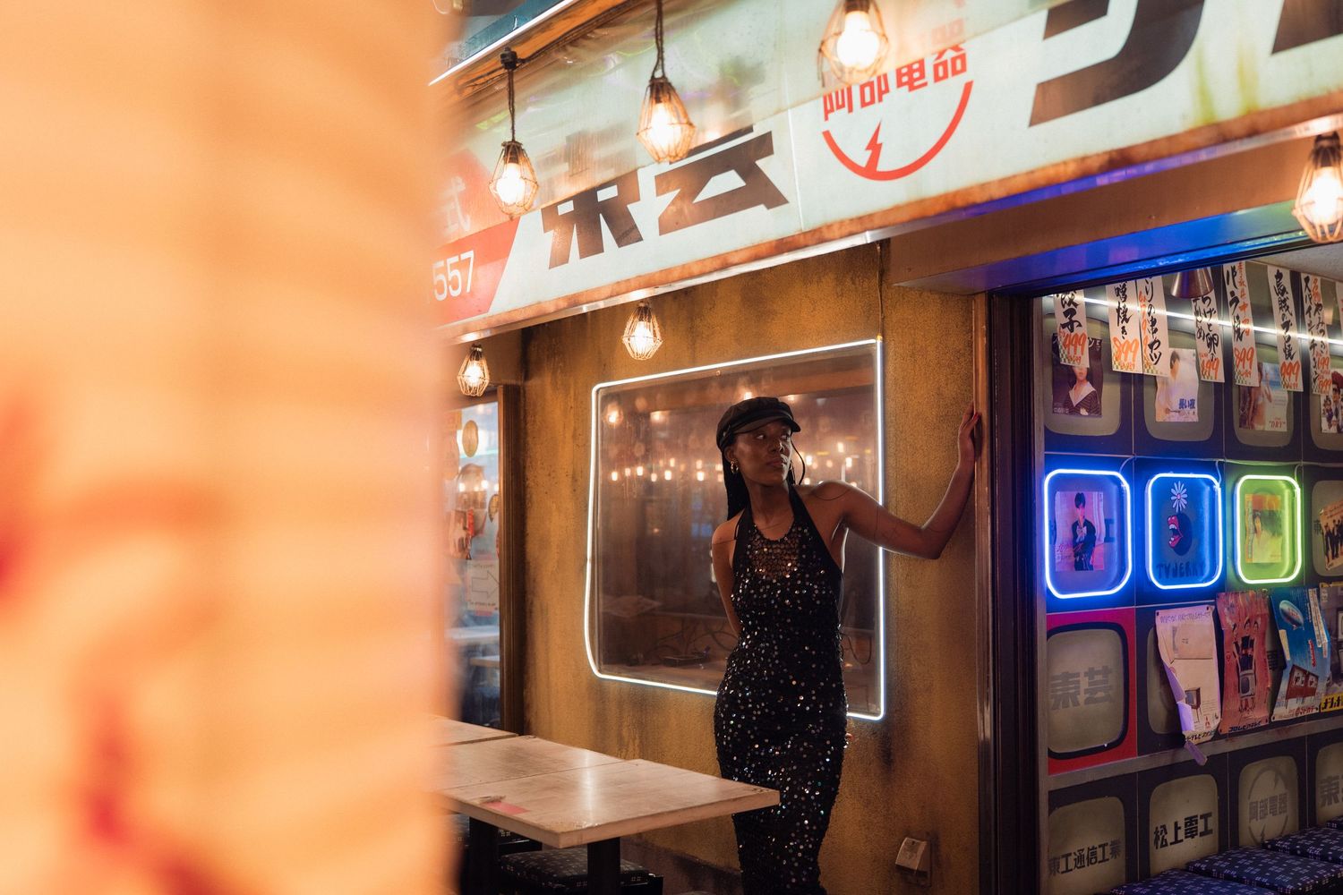 Woman in sequin dress poses by neon-lit Asian restaurant storefront at night.