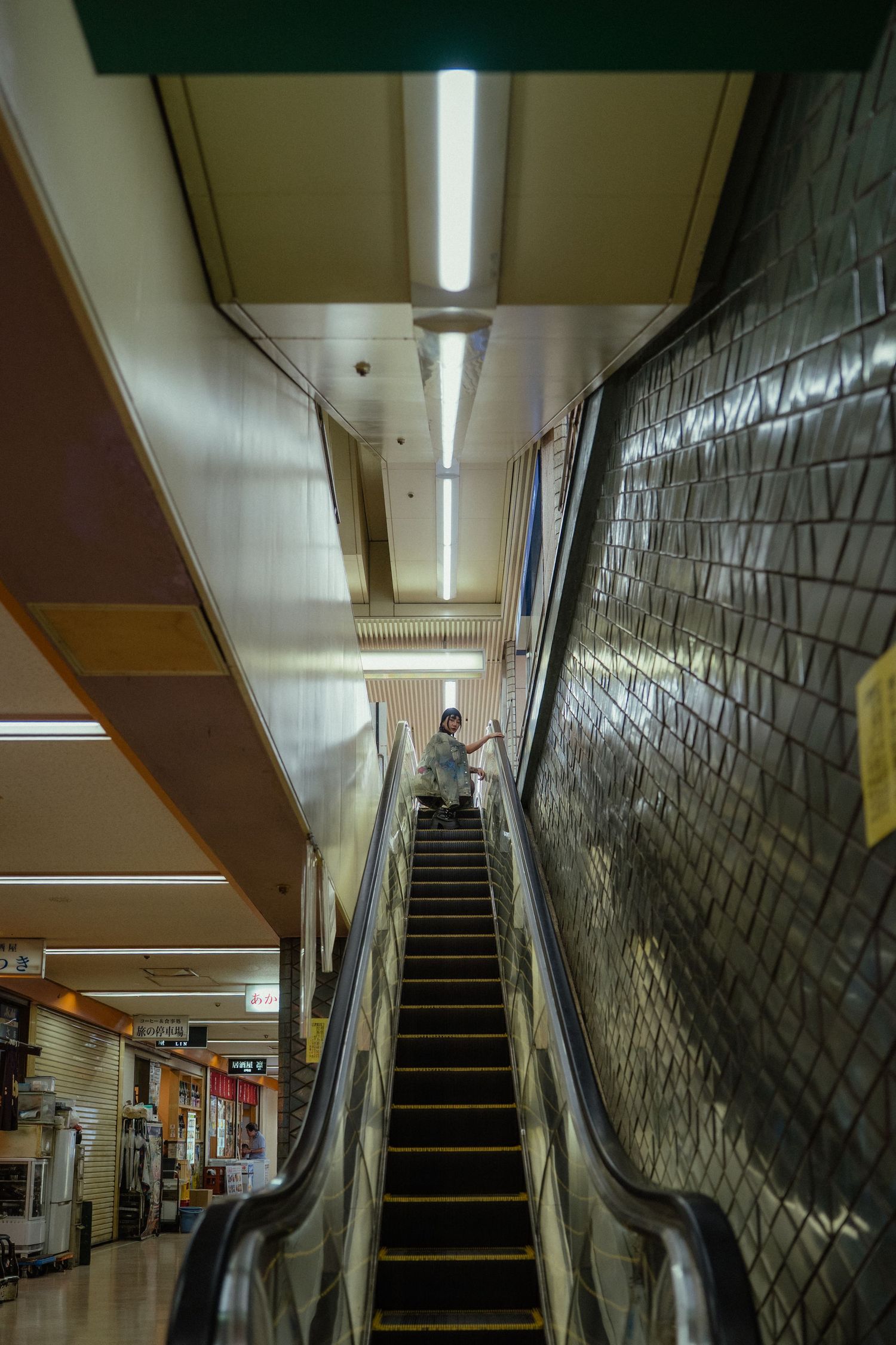 Person riding an escalator in a subway station with tiled walls and fluorescent lighting overhead.
