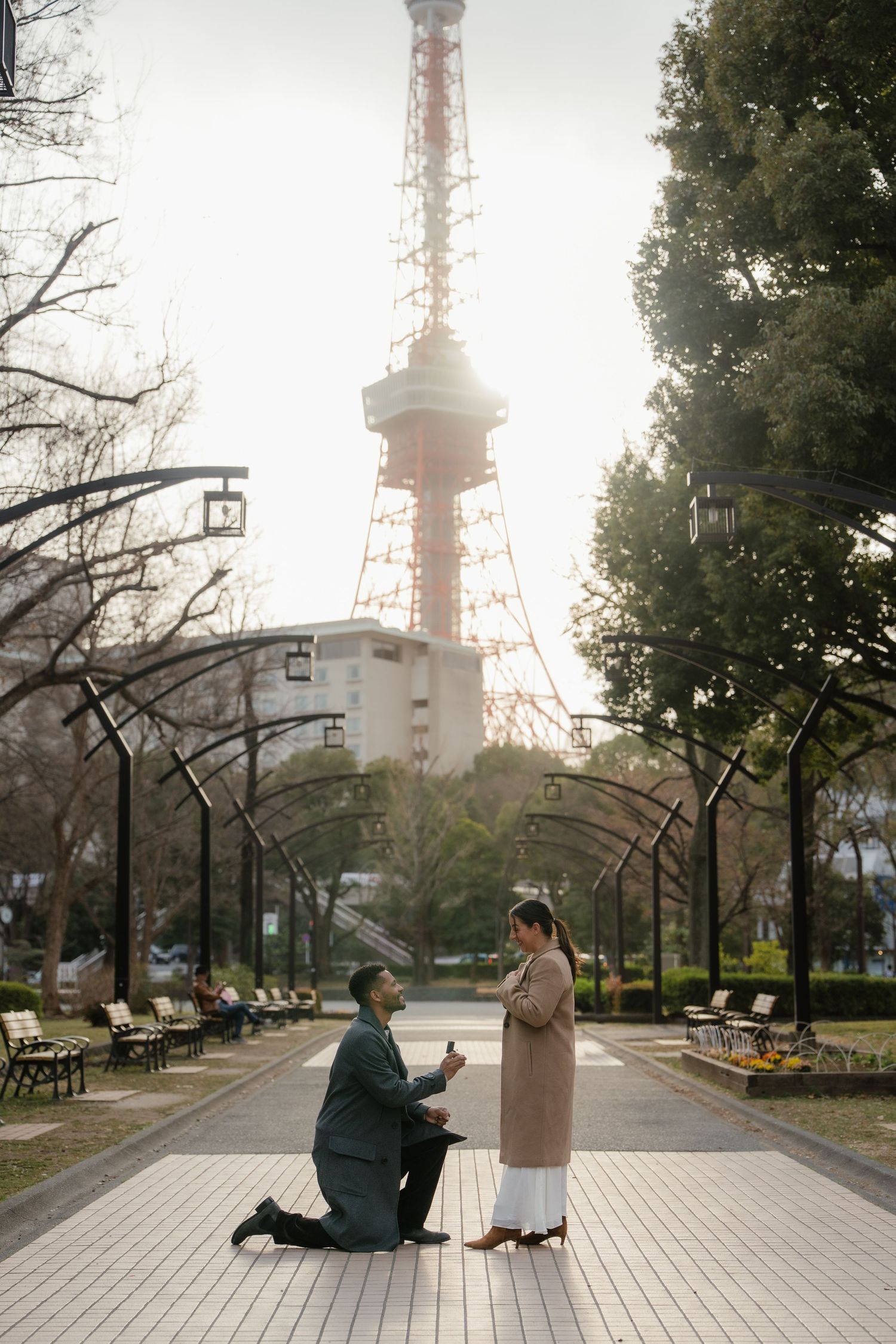 Man proposes on one knee to woman in park with Tokyo Tower glowing in background at sunset.