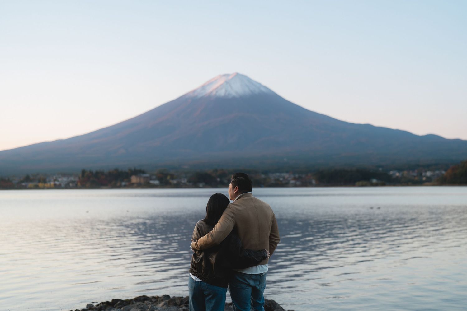Couple embracing by a serene lake with iconic snow-capped Mount Fuji in the background at dusk.