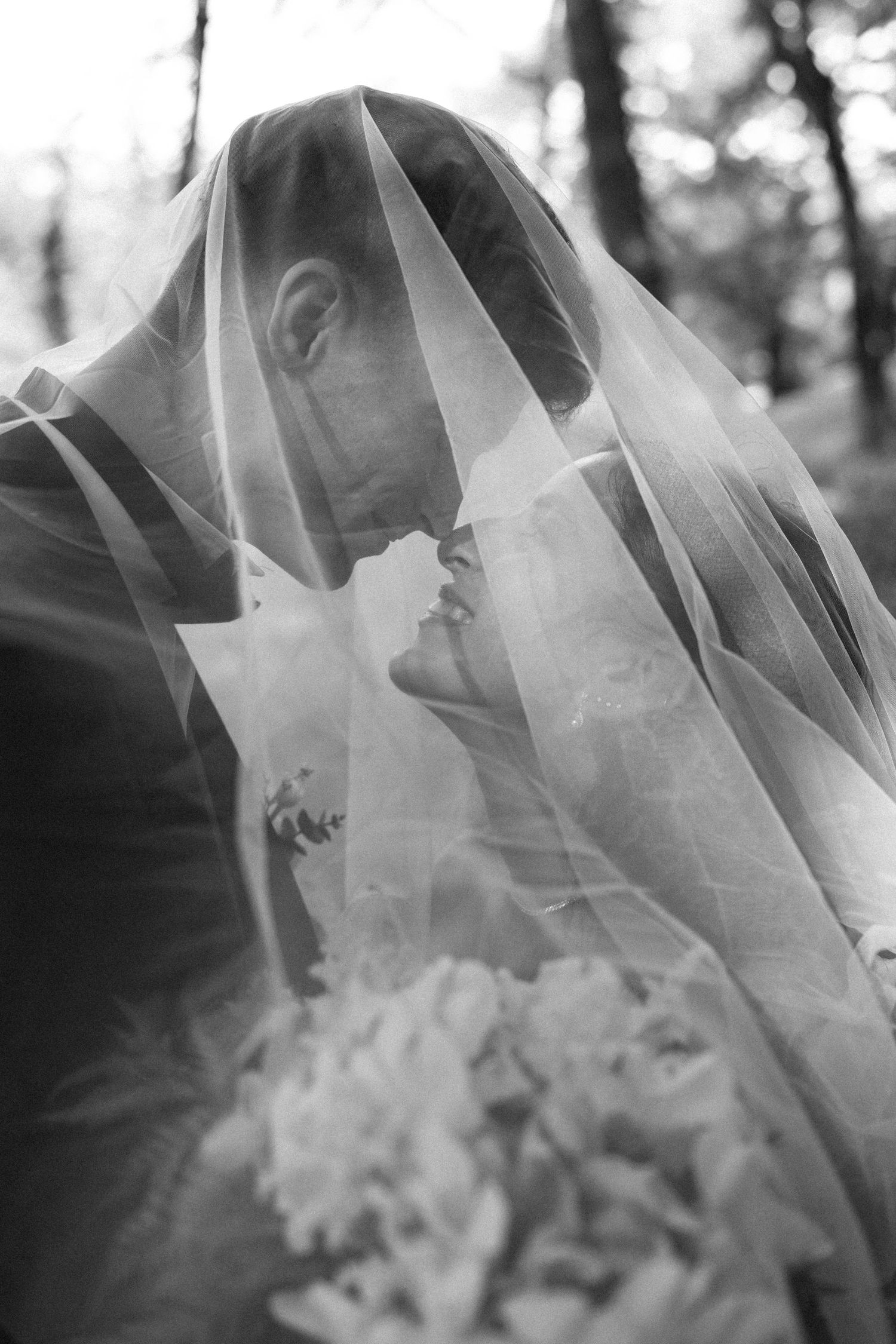 Black and white photo of bride and groom sharing a kiss under a flowing wedding veil in a forest setting.