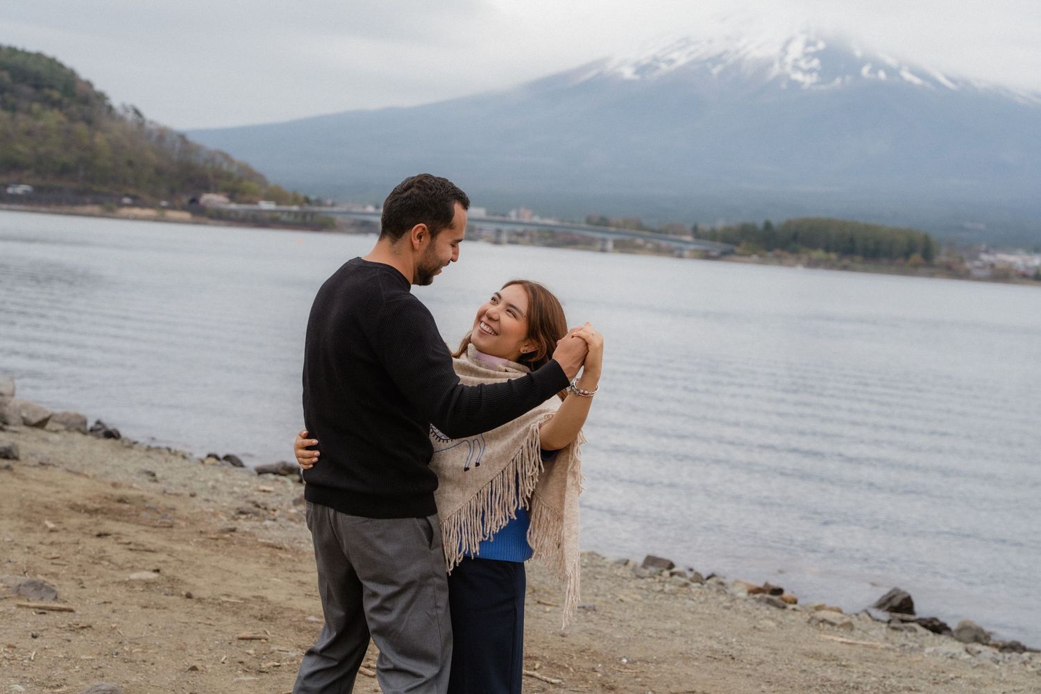 Couple embracing on lakeside beach with snow-capped Mount Fuji in background on cloudy day in Japan.