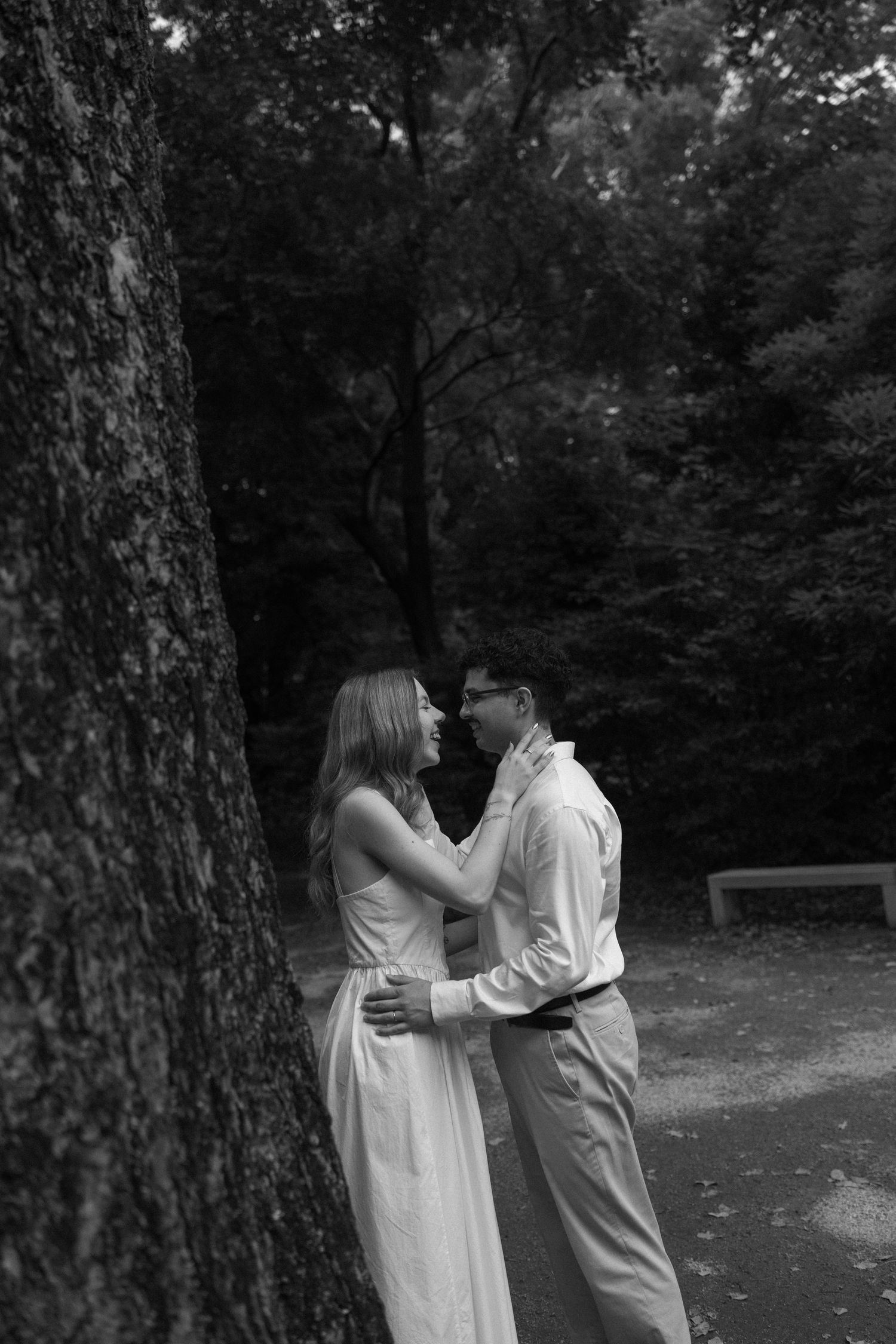 Black and white photo of a romantic couple embracing near a large tree in a lush park setting.