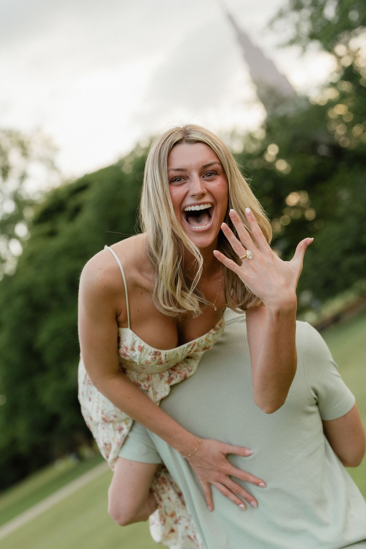 Excited blonde woman showing off engagement ring while being carried piggyback in a lush green park setting.