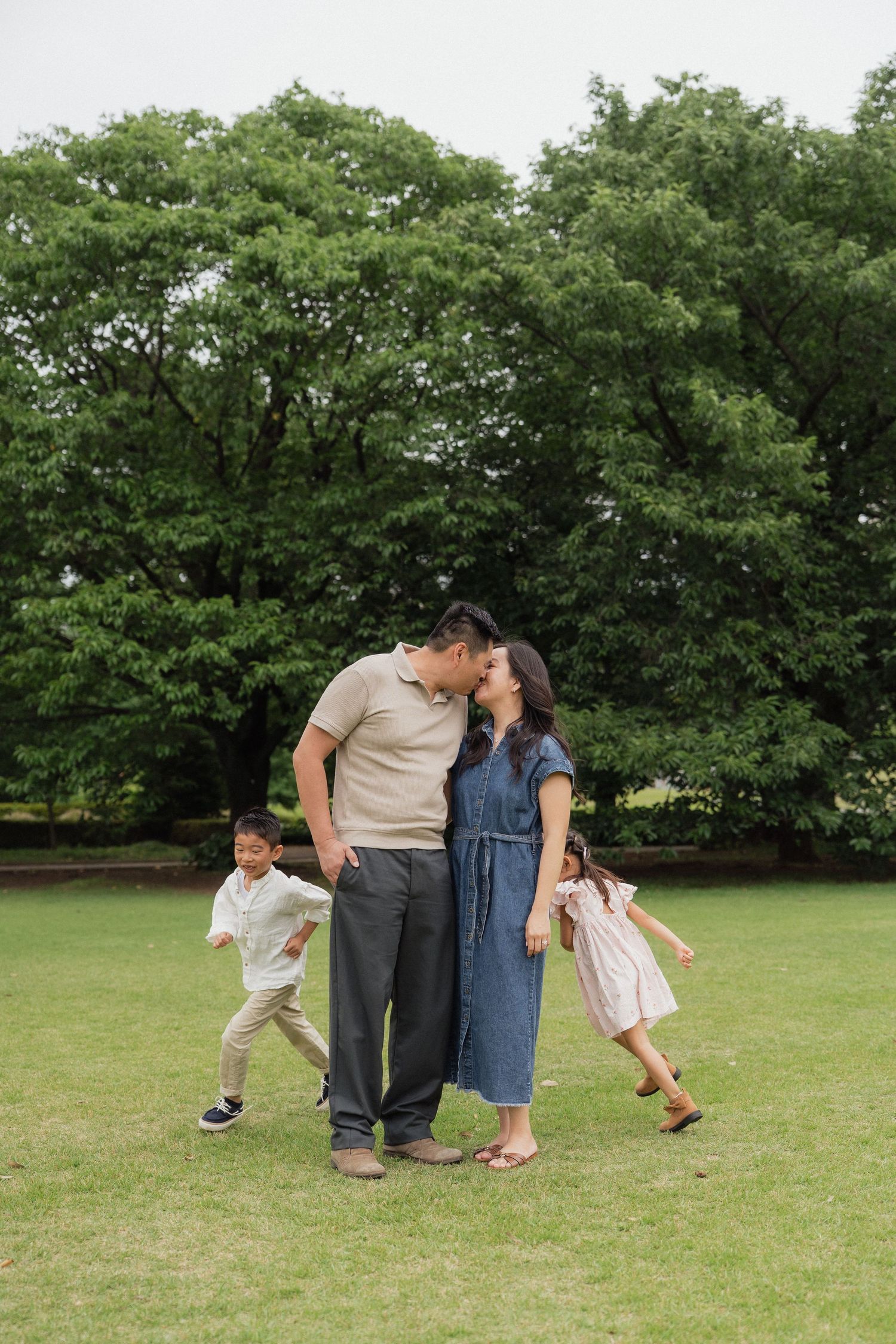Family of four enjoying a sunny day in the park, parents sharing a kiss while children play around them.