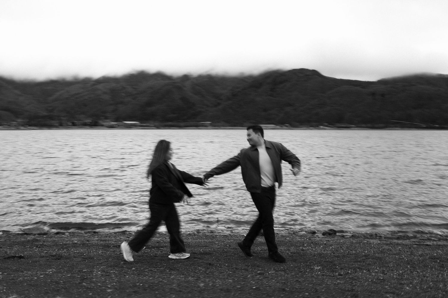 Black and white photo of a couple holding hands walking along a rocky shoreline with misty mountains in the background.