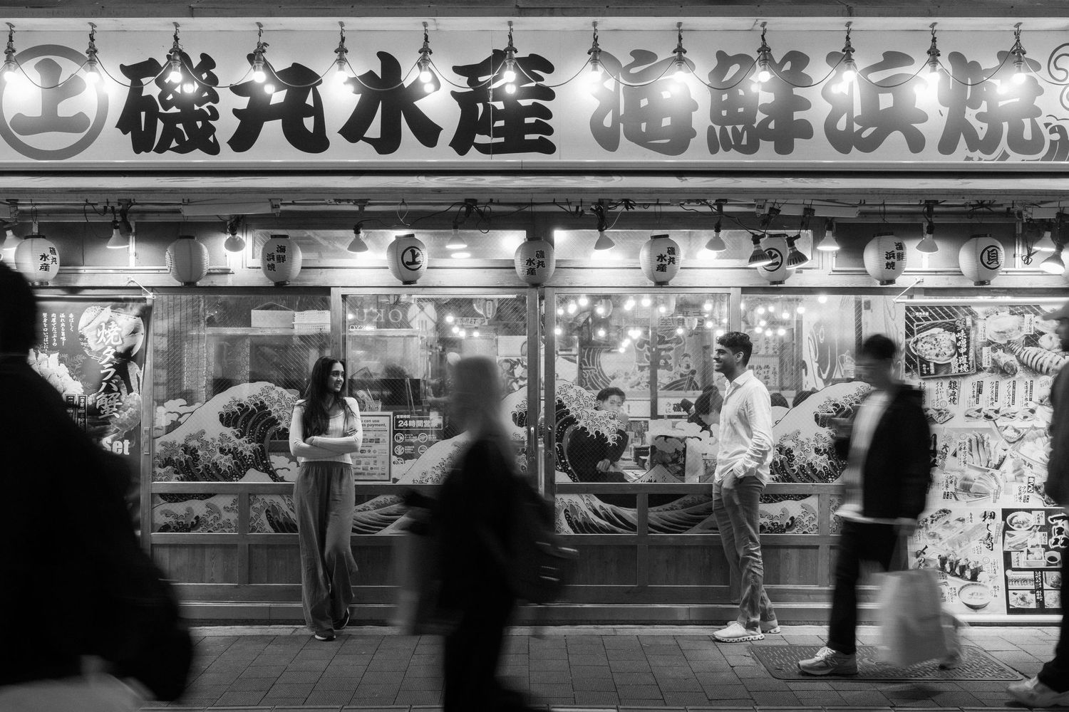 Black and white photo of a bustling Japanese seafood restaurant storefront at night with illuminated lanterns and kanji signage.