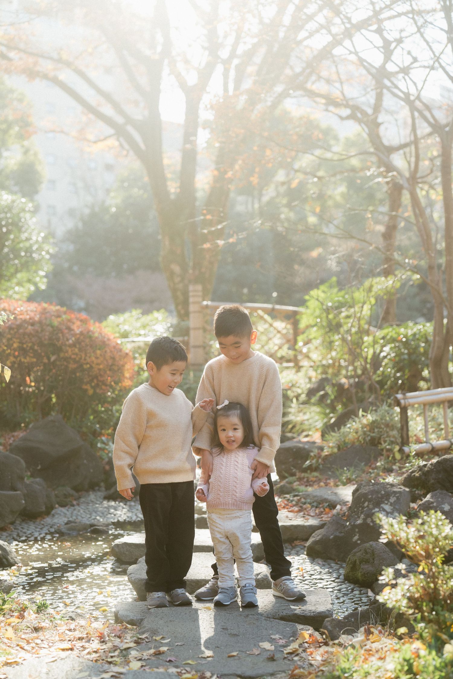 Three children stand on stepping stones in a sunlit Japanese garden surrounded by autumn foliage.