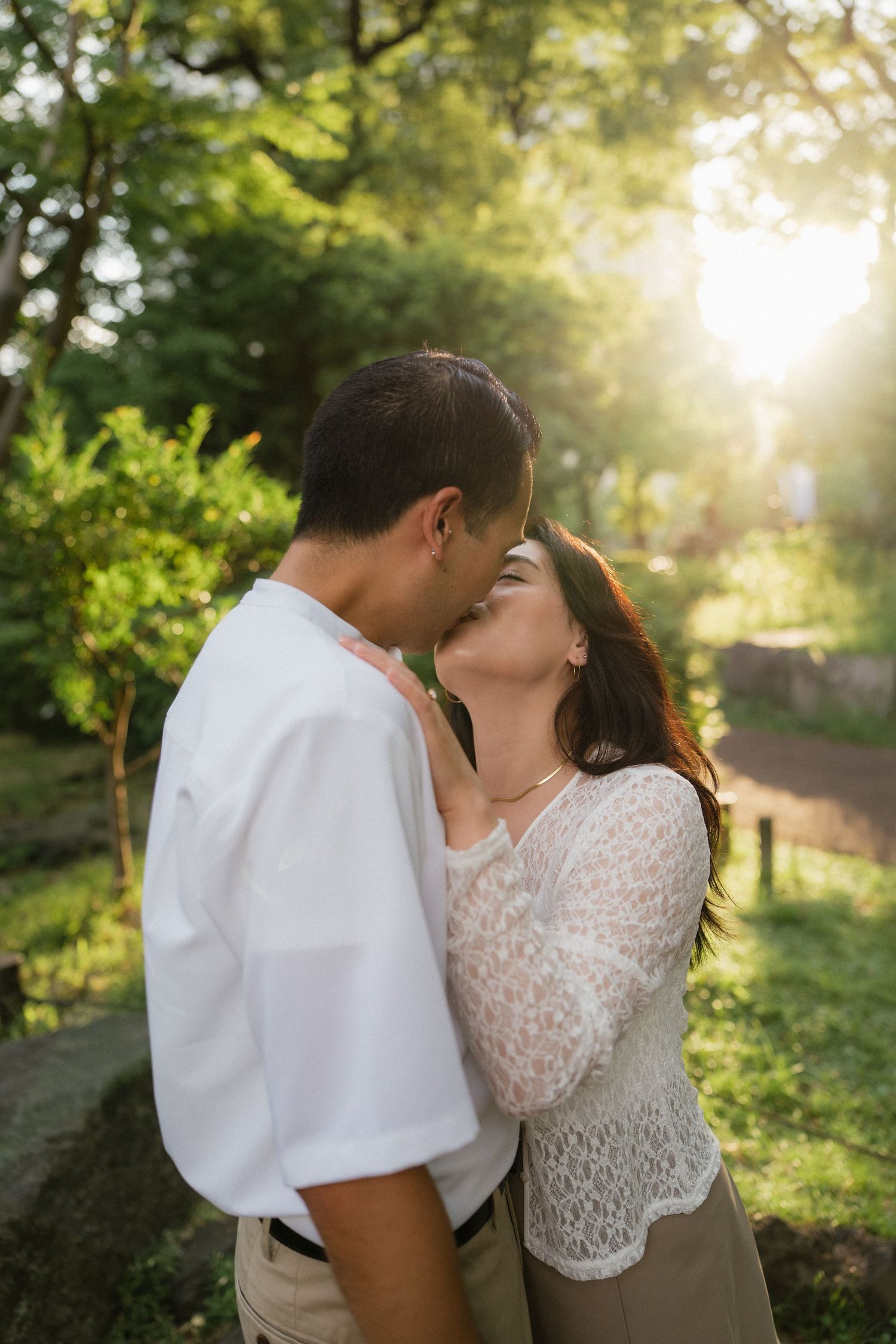 Couple sharing a romantic kiss outdoors in a sunlit green park, man in white shirt and woman in lace top.