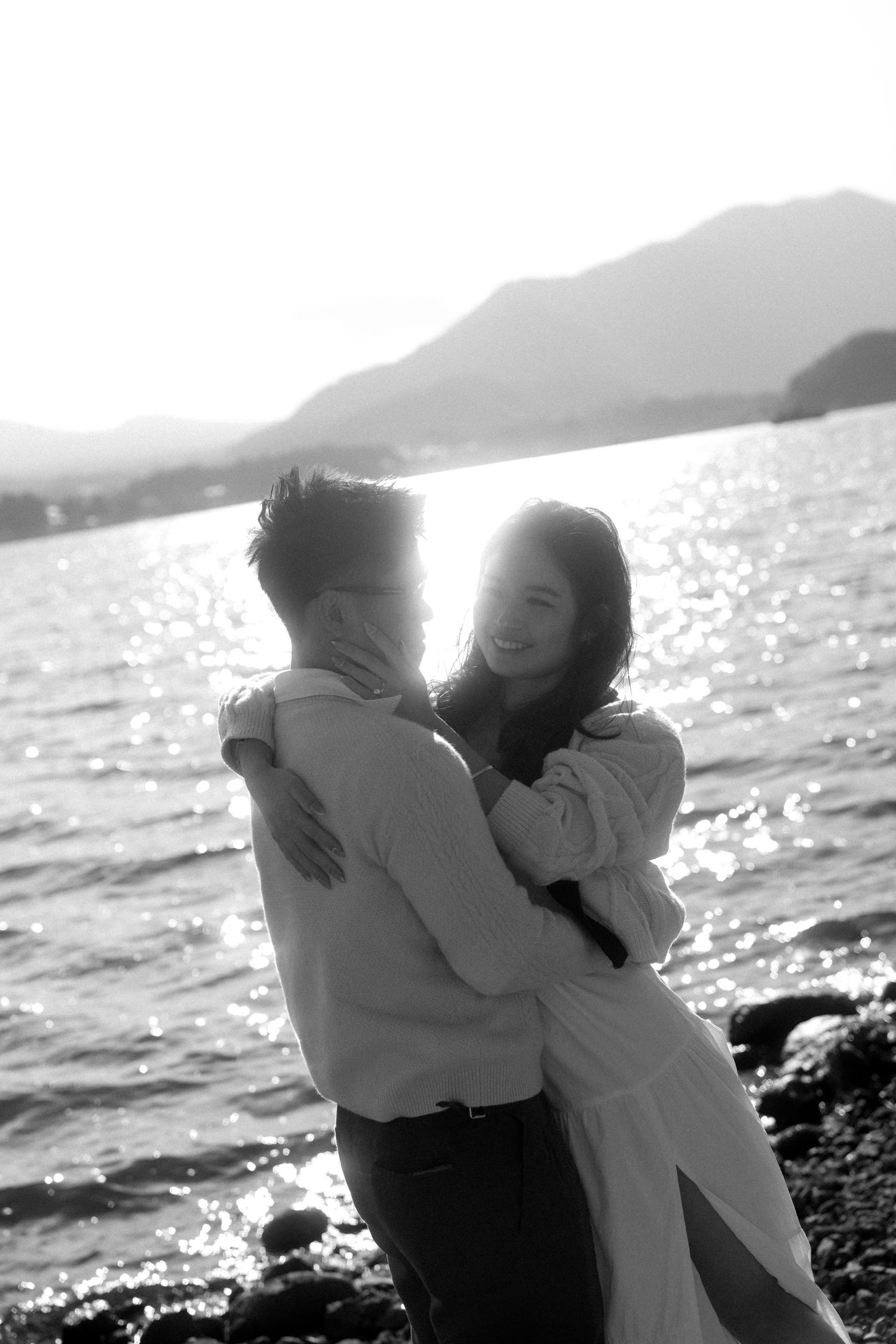 Black and white photo of a couple embracing romantically by a shimmering lake with mountains in the background.