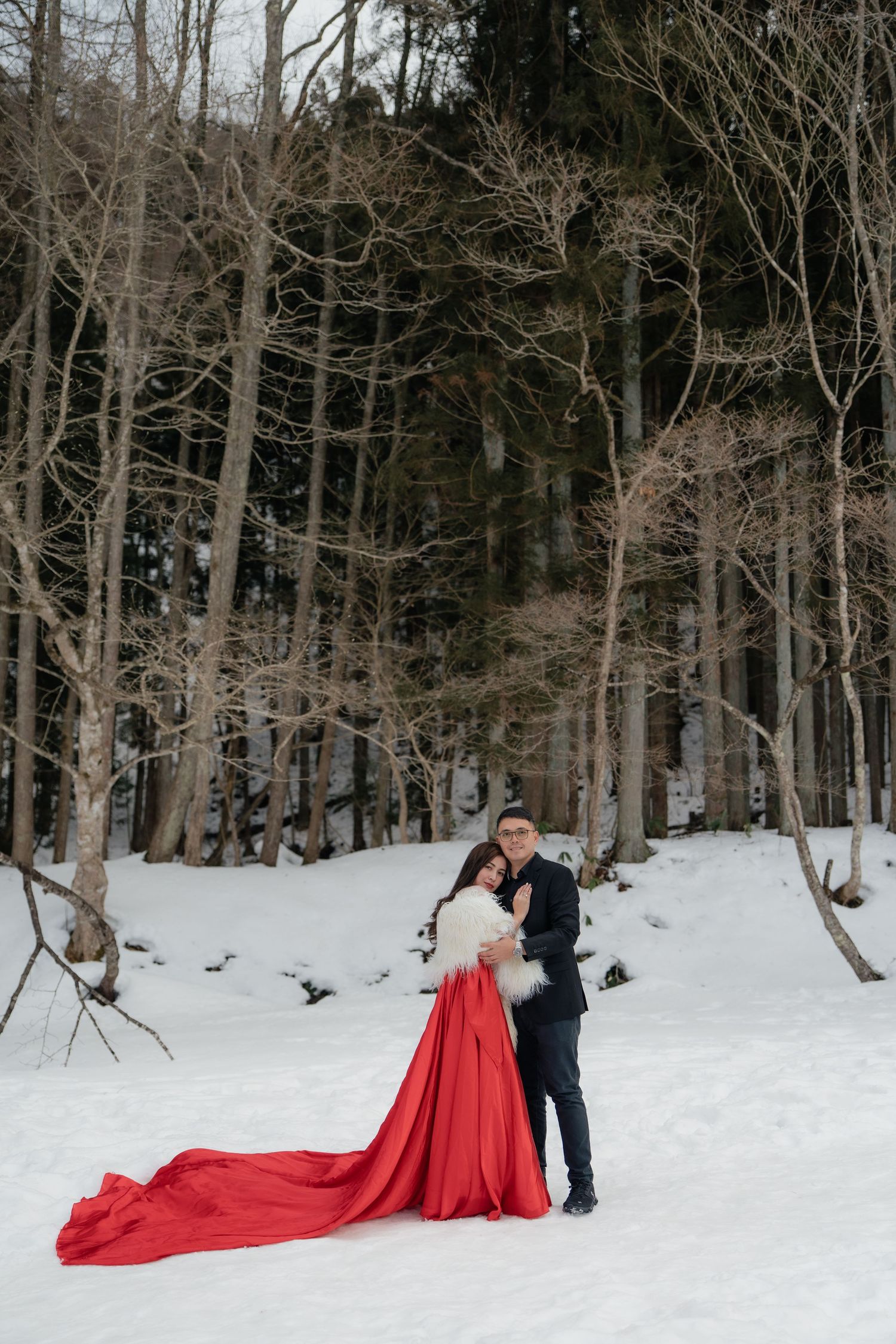 Couple poses in snowy winter forest, bride in red gown with long train, groom in navy suit embracing her.