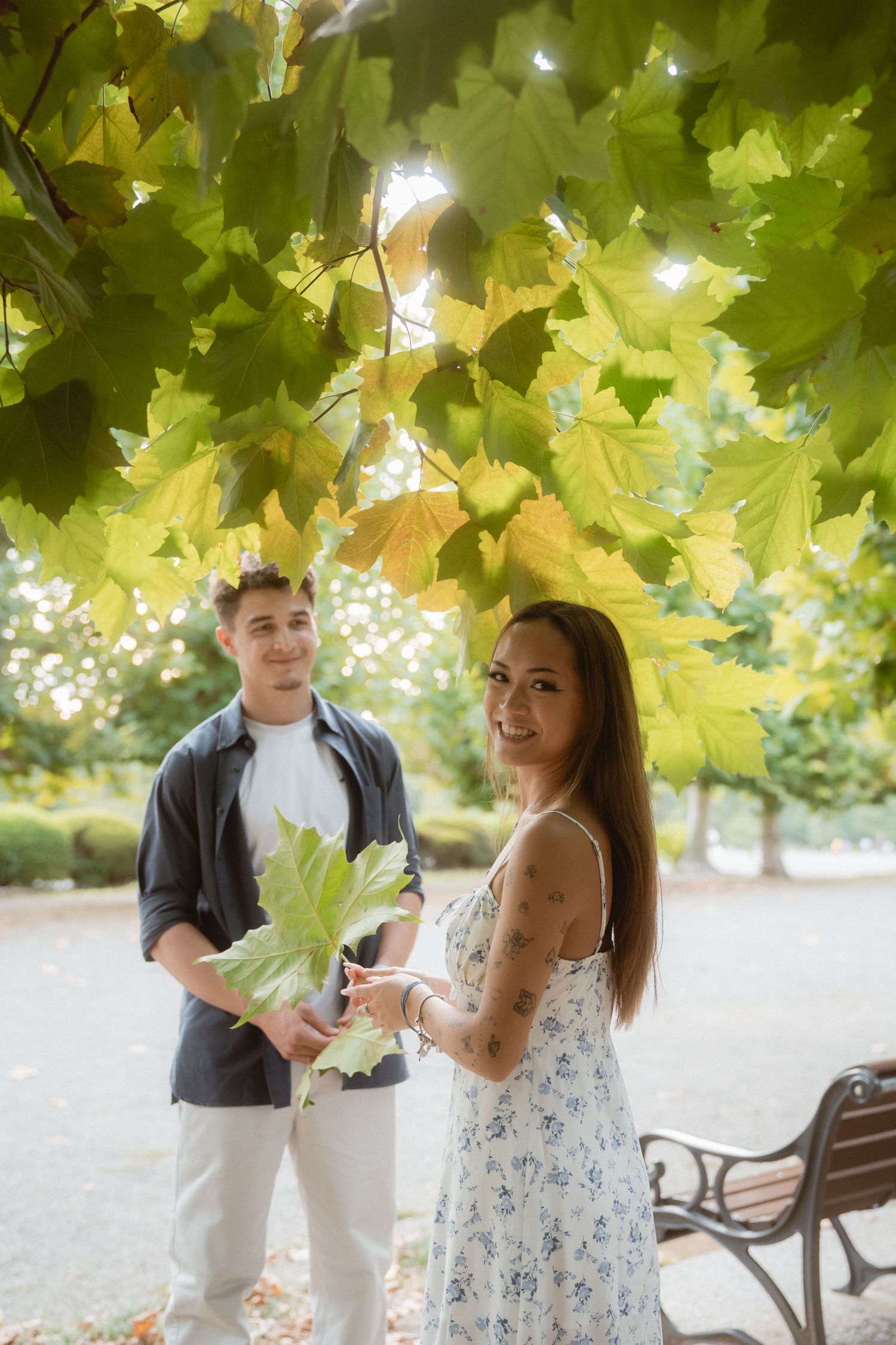 A young couple stands under maple trees in a park, the woman smiling while the man holds a large green leaf.