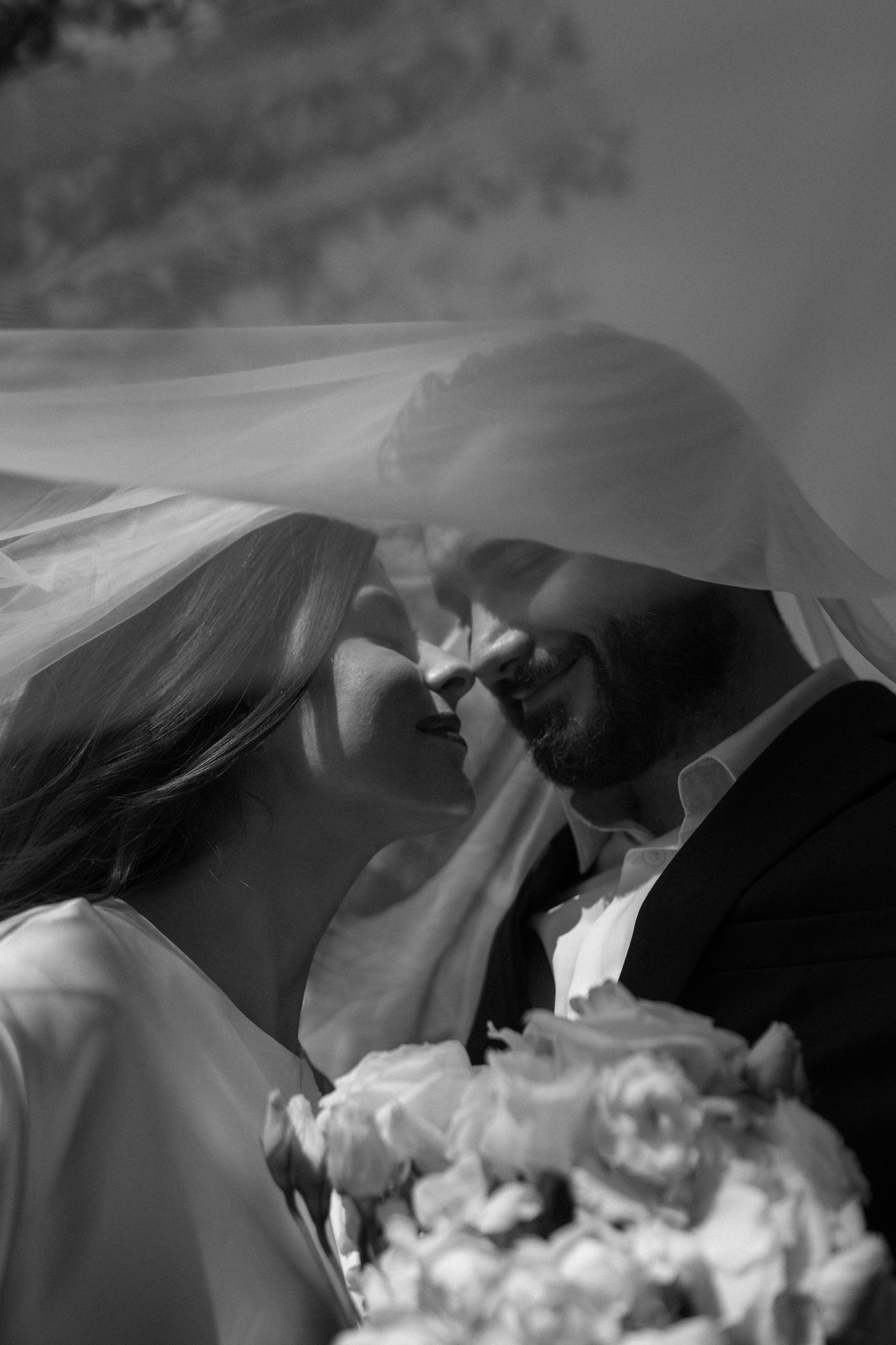Black and white wedding photo of bride and groom sharing intimate moment under veil, bride holding bouquet.