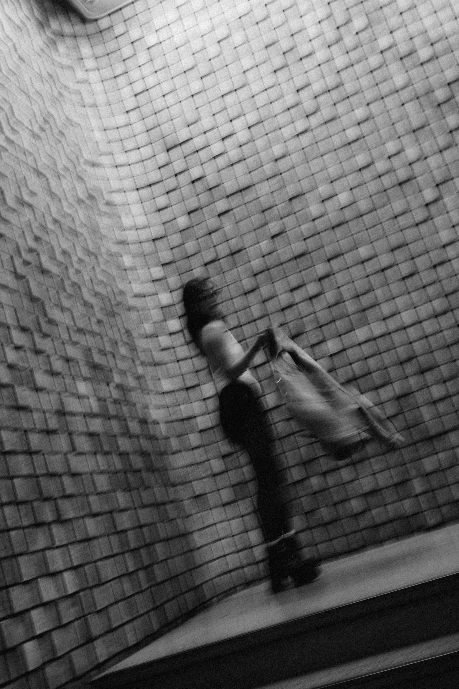 Blurred black and white photo of a woman walking past a textured woven tile wall, creating motion effect.