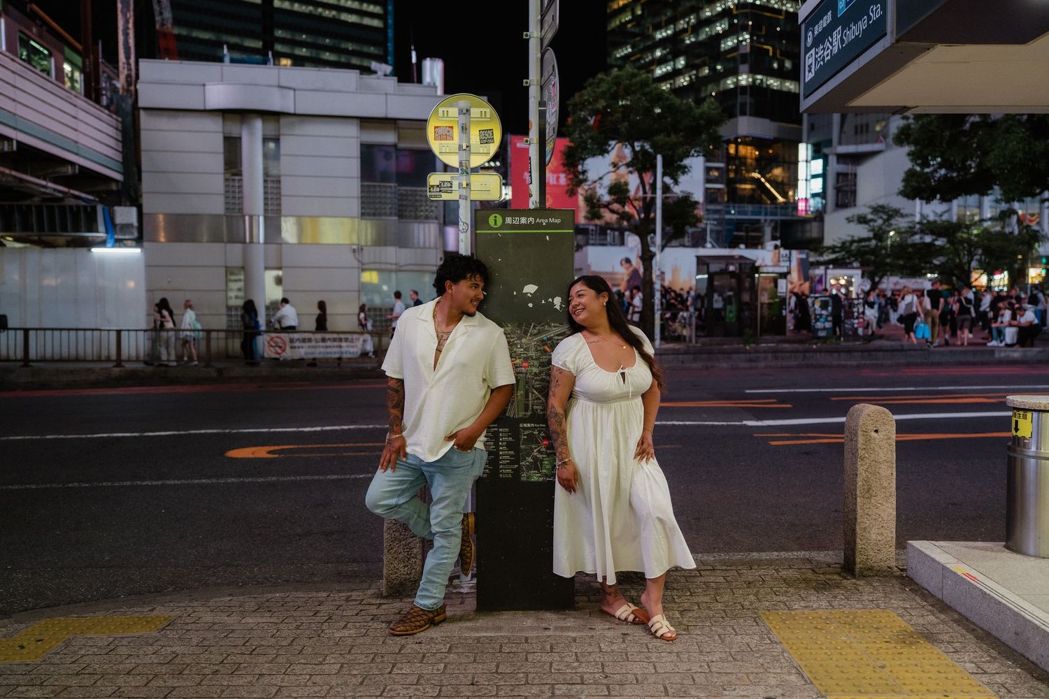 Couple standing by a bus stop sign at night on a busy urban street in Japan.