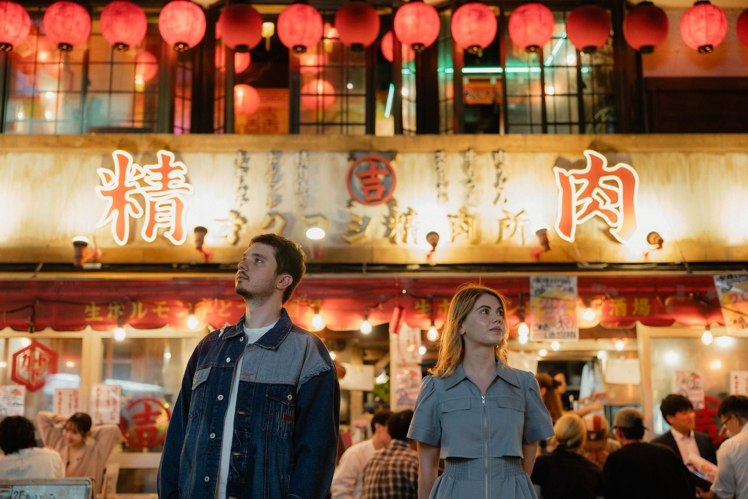 Couple gazing up at glowing red lanterns outside busy Asian night market restaurant.