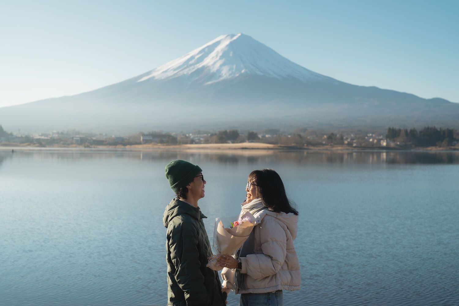 Couple holding hands by Lake Kawaguchiko with iconic snow-capped Mount Fuji reflected in calm waters on a clear day.