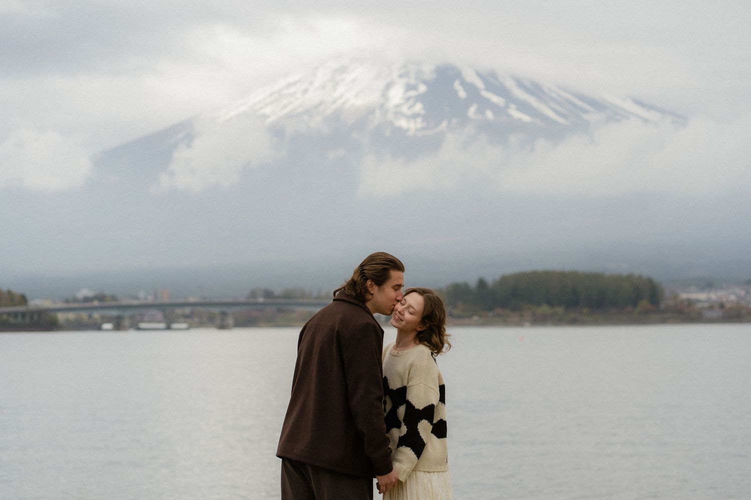 Couple sharing a romantic moment by a lake with Mount Fuji in the background on a cloudy day.