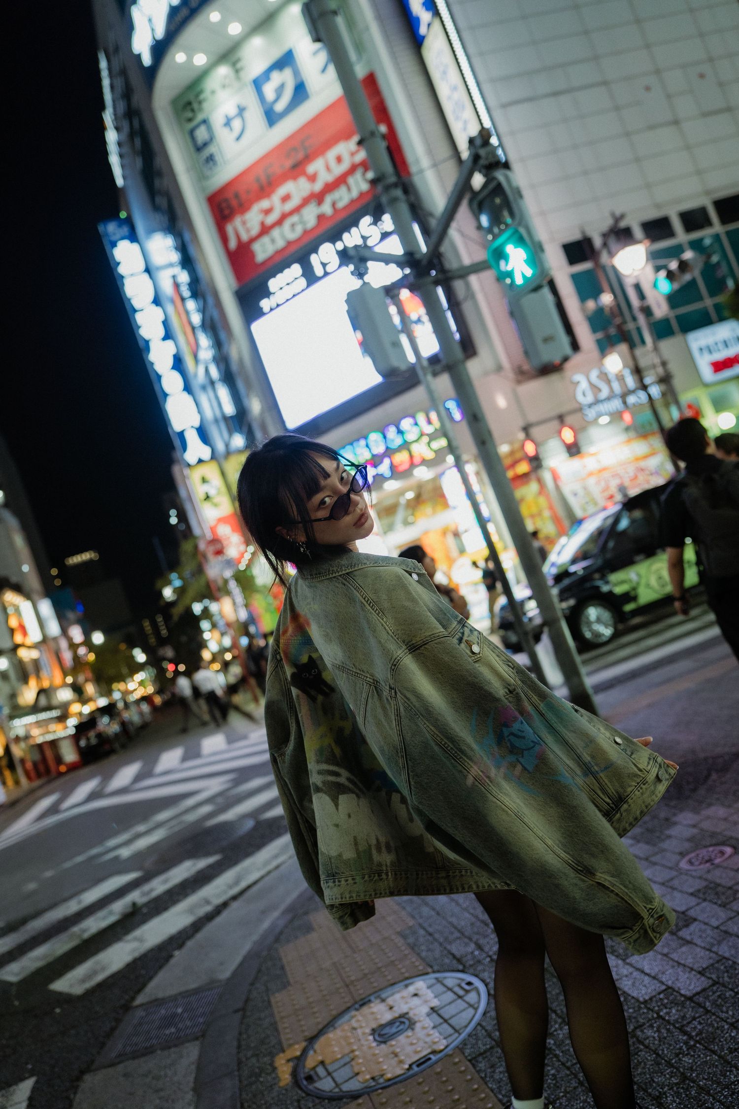 Young woman in oversized green jacket smiling on a vibrant Tokyo street at night with neon signs.