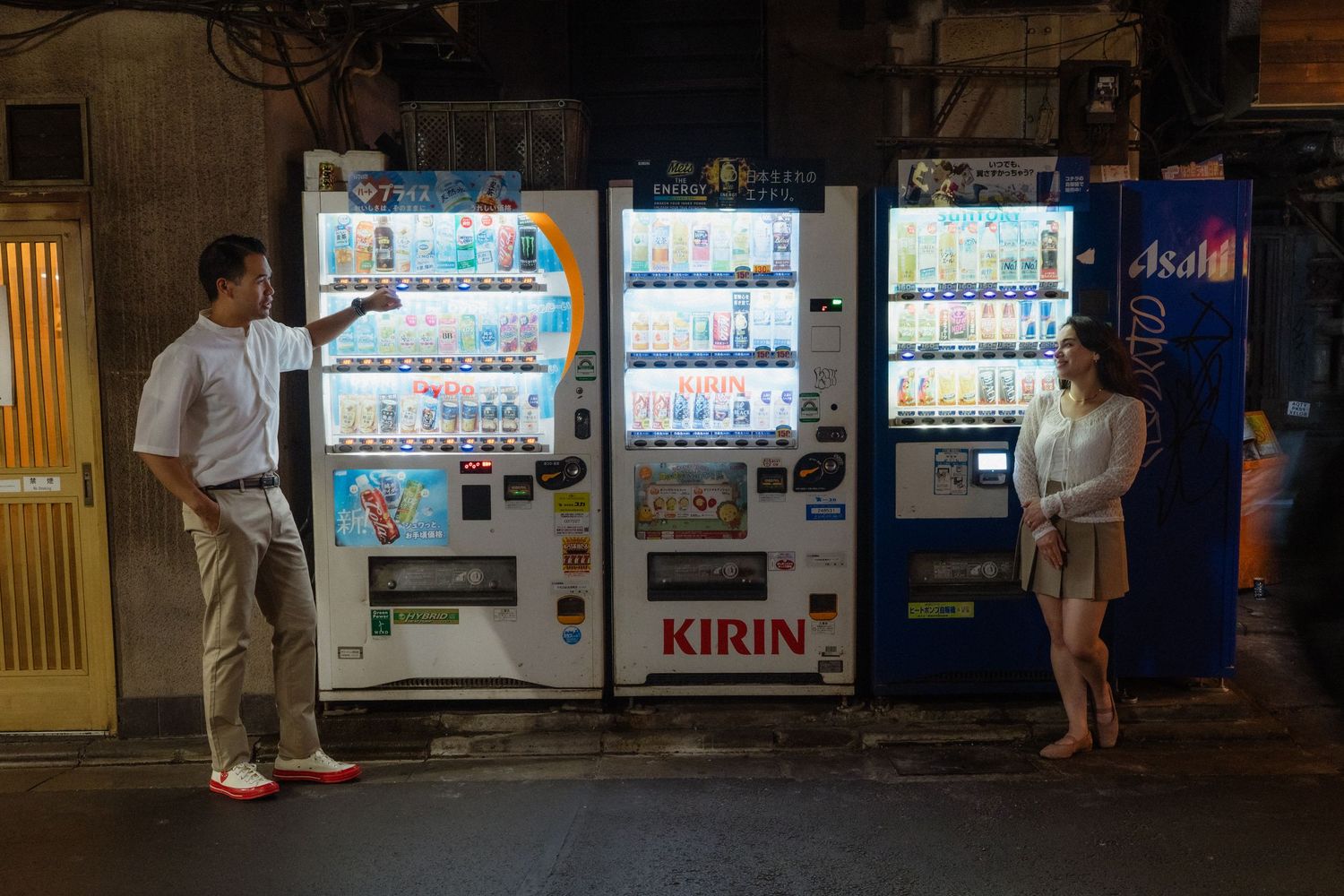 Two people browsing colorful Kirin and Asahi vending machines on a Japanese street at night.