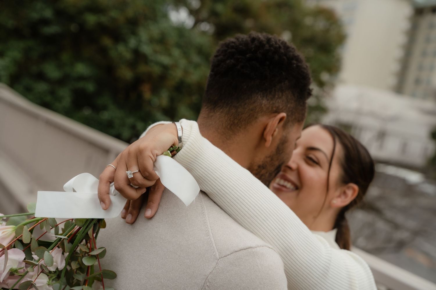 A joyful couple embraces outdoors, the woman smiling warmly while holding a bouquet, dressed in elegant white attire.