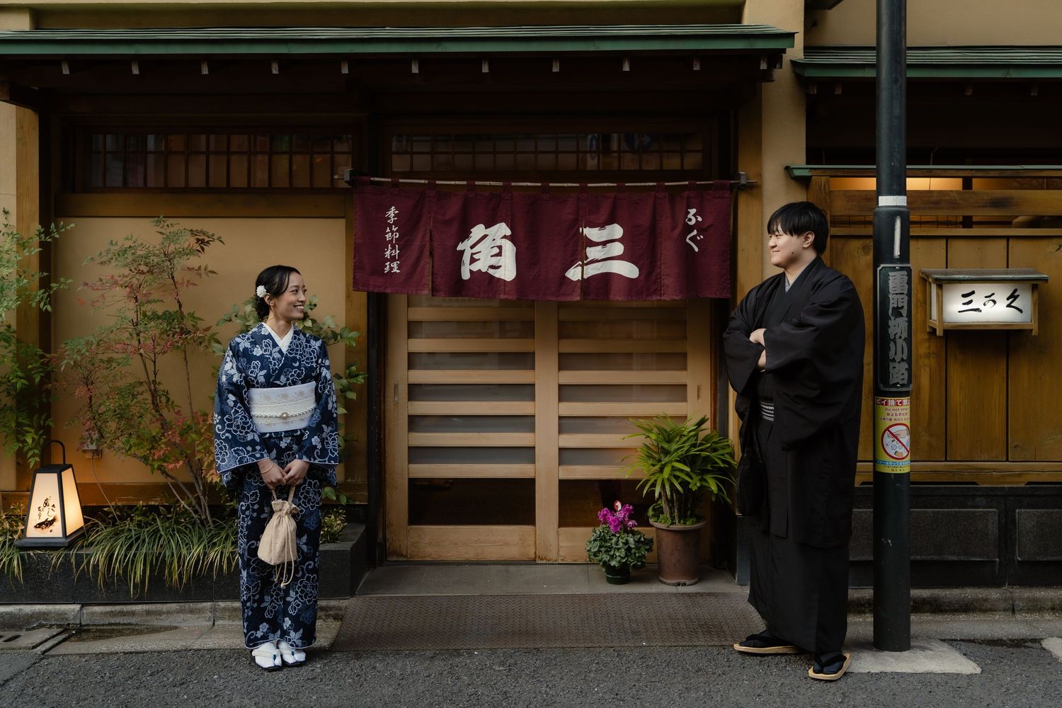 Two people in traditional Japanese kimono stand outside a restaurant with a red noren curtain in Kyoto, Japan.