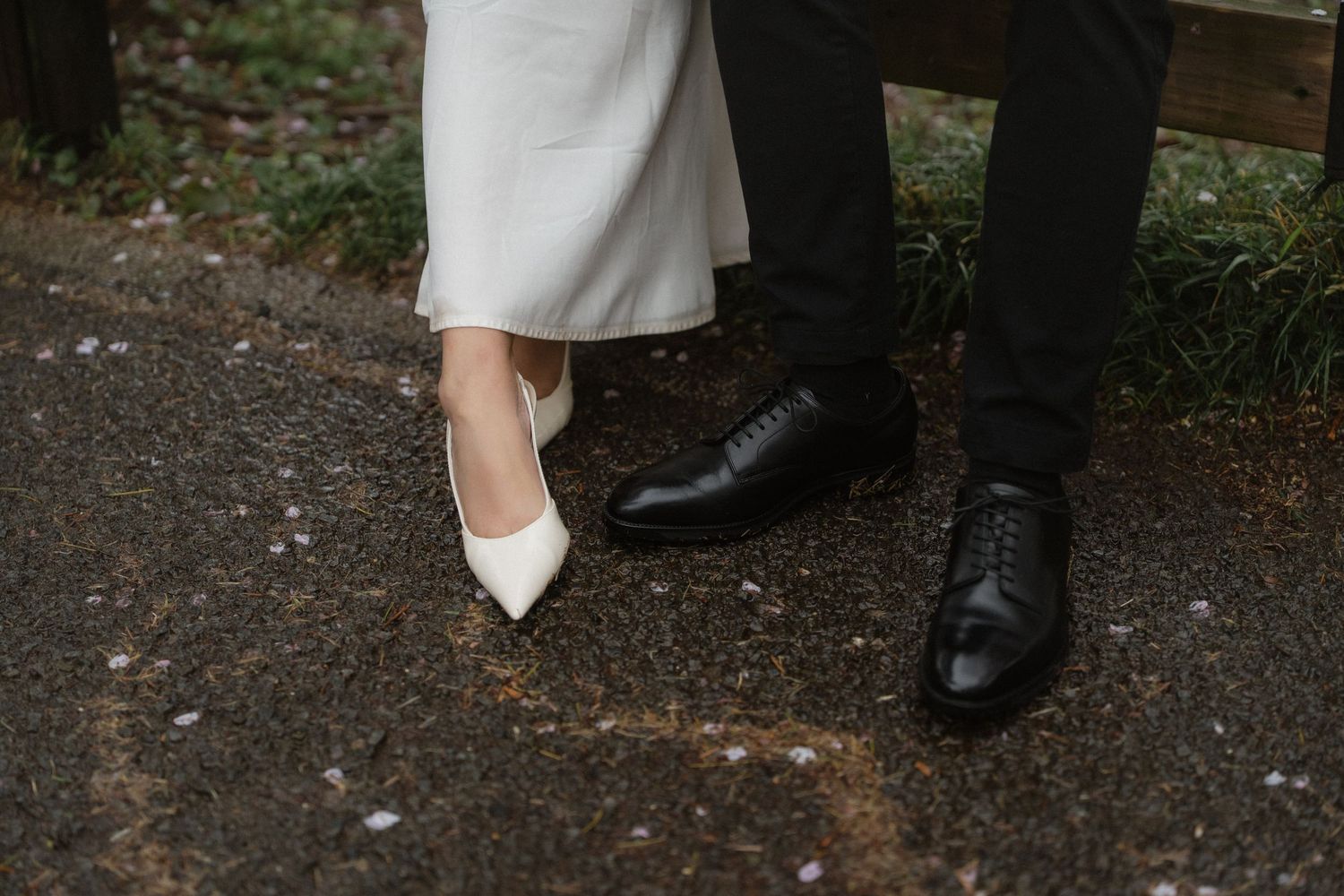 Close-up of wedding couple's feet, bride in white heels and groom in black dress shoes on a paved path.