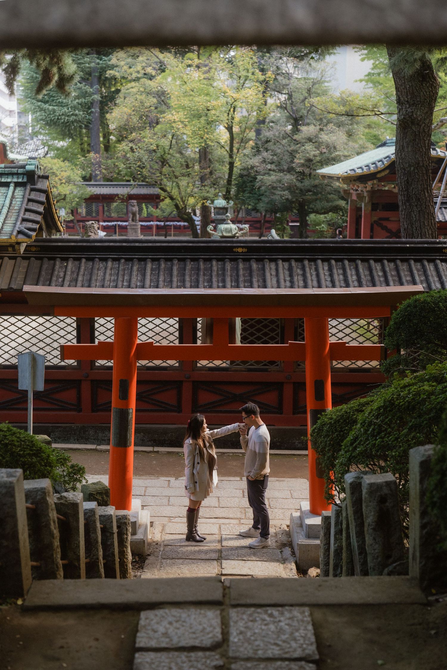 Couple standing beneath a vibrant red torii gate at a Japanese shrine surrounded by lush greenery.