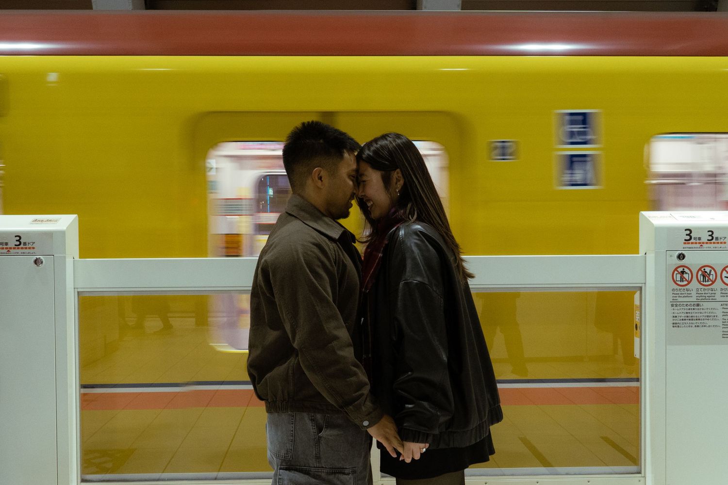 Couple sharing a romantic kiss on a subway platform with a yellow train passing behind them.