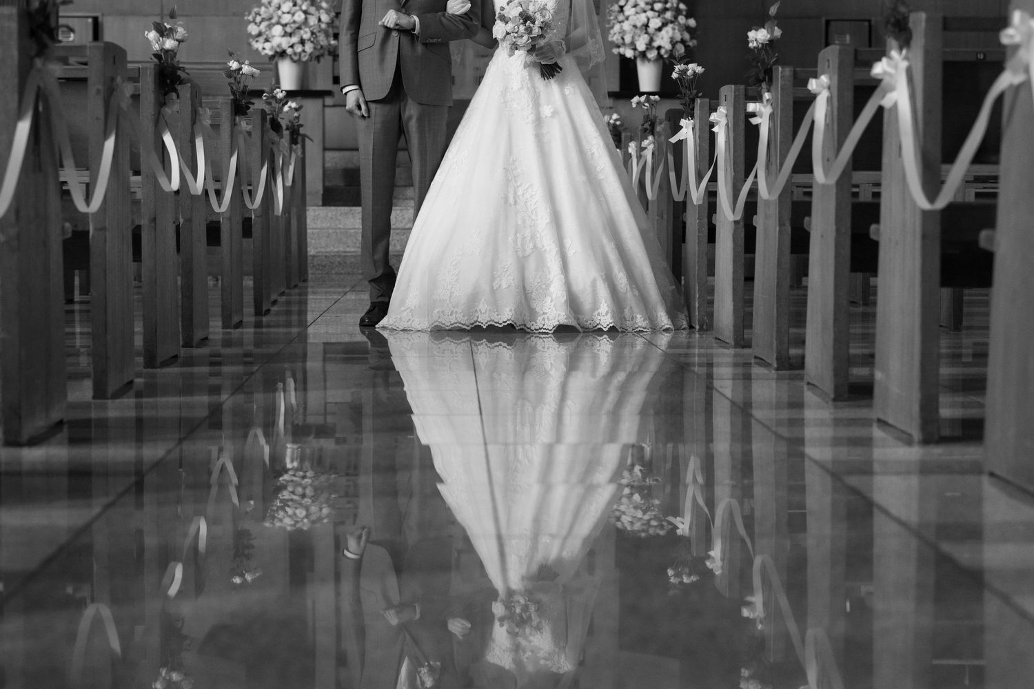 Black and white photo of a bride walking down a church aisle, her gown reflected on the polished marble floor.