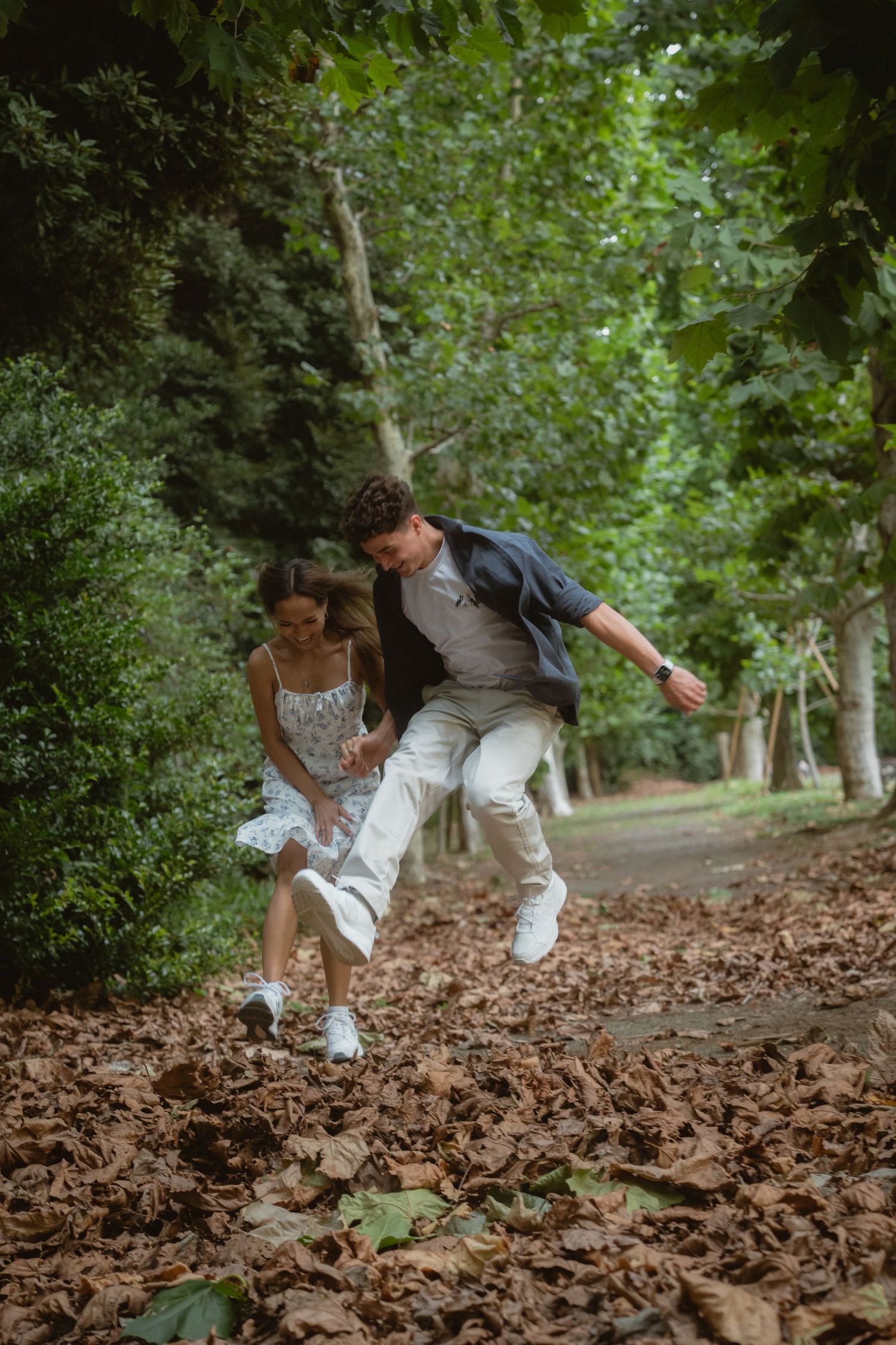 A couple joyfully running through a leaf-covered park path surrounded by lush green trees.