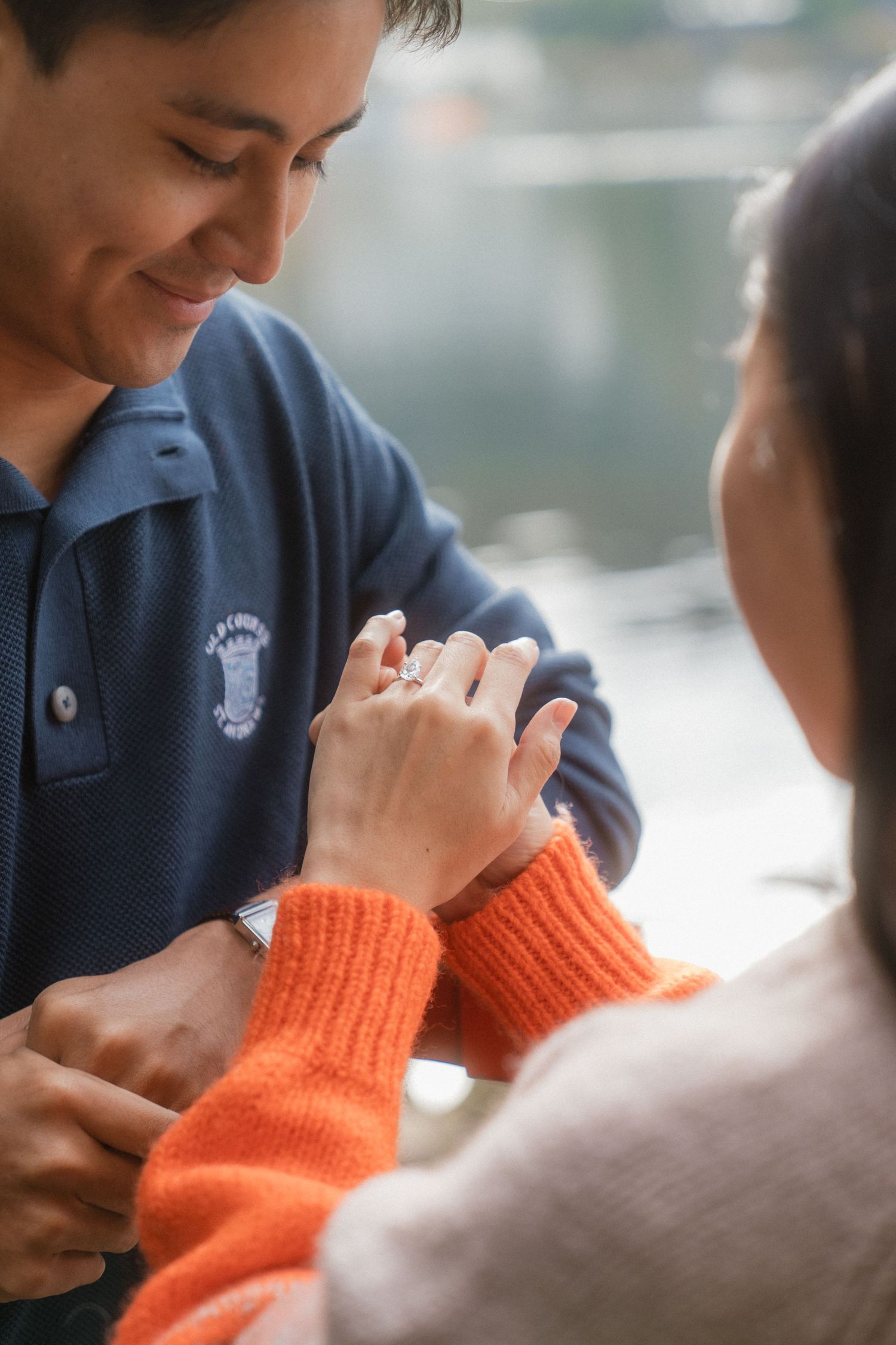 A smiling man in a blue polo shirt places a diamond engagement ring on a woman's finger in a romantic proposal moment.