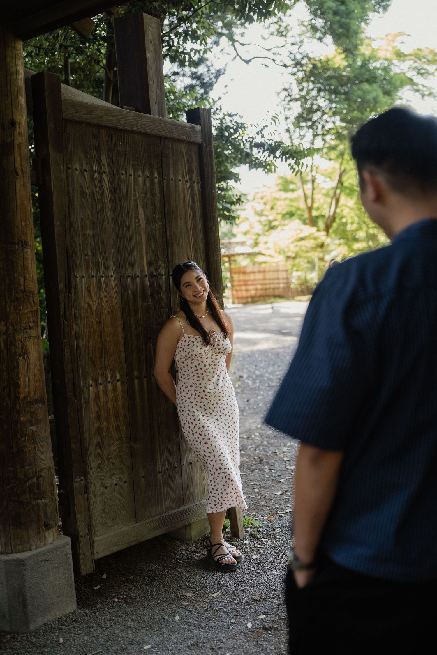 Woman in polka dot dress leaning against wooden gate smiling at man in Japanese garden setting.