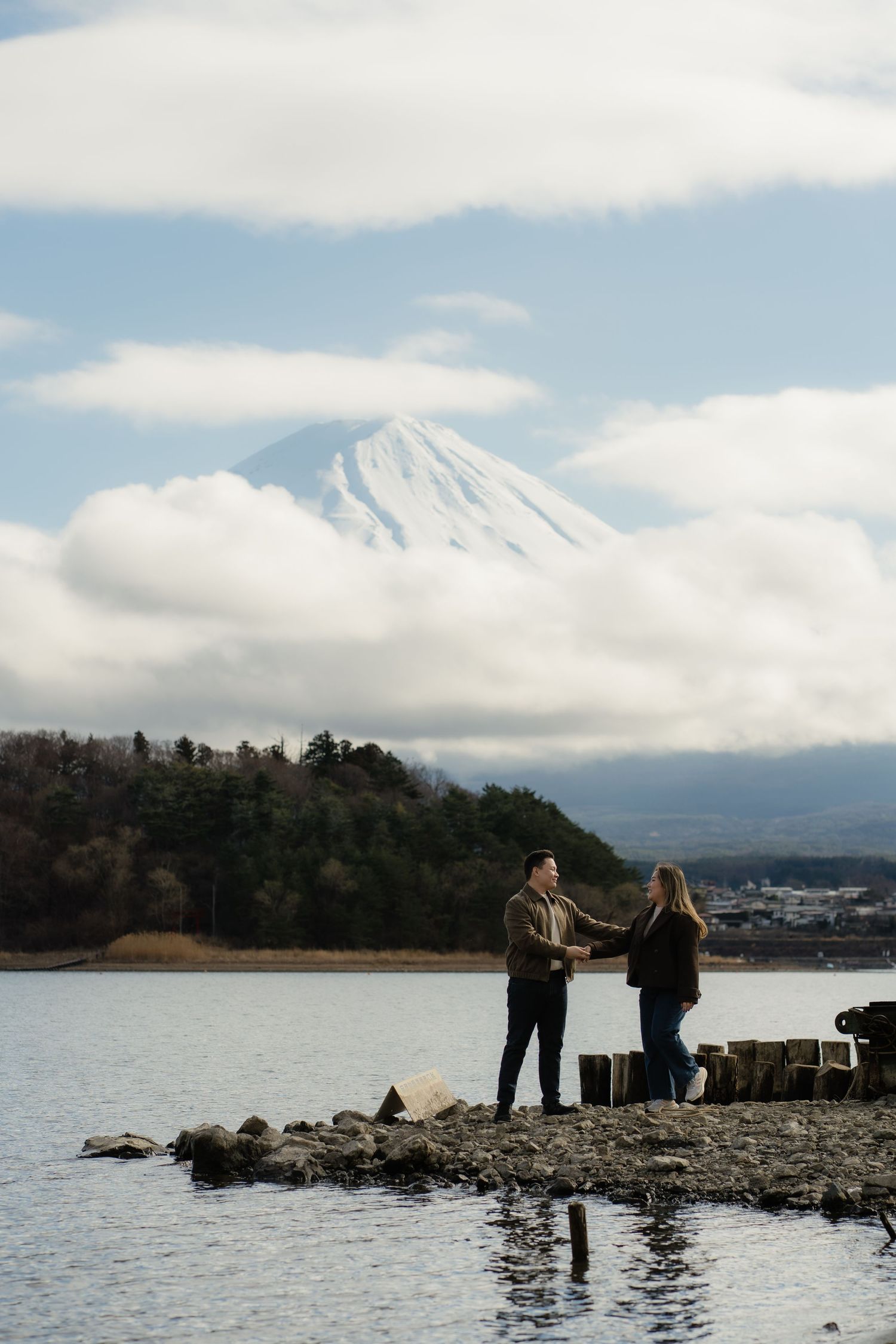 Couple holding hands on rocky shore with snow-capped Mount Fuji and clouds in background, Japan.