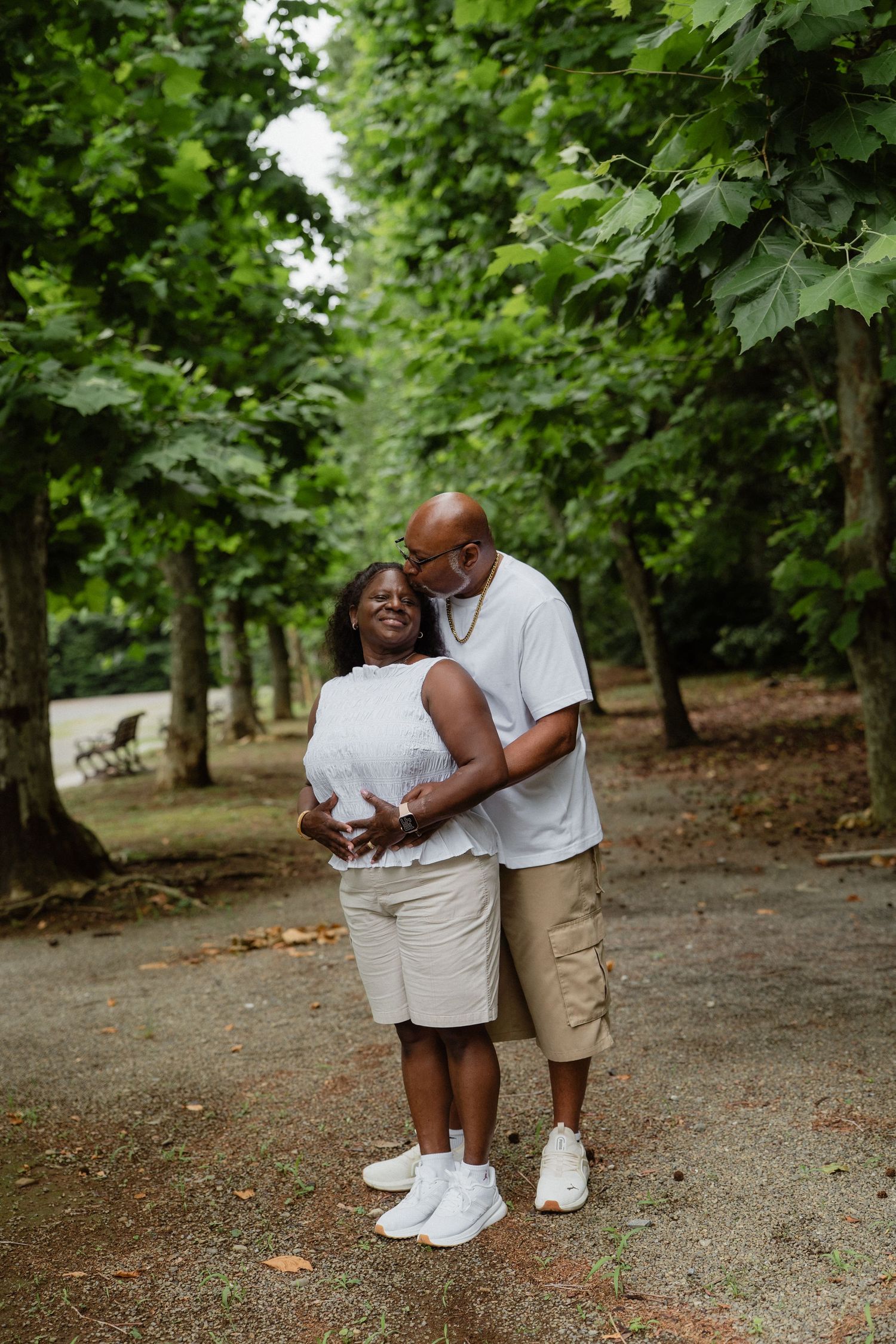 A couple embraces warmly on a tree-lined path in a lush green park, both wearing white tops and casual shorts.