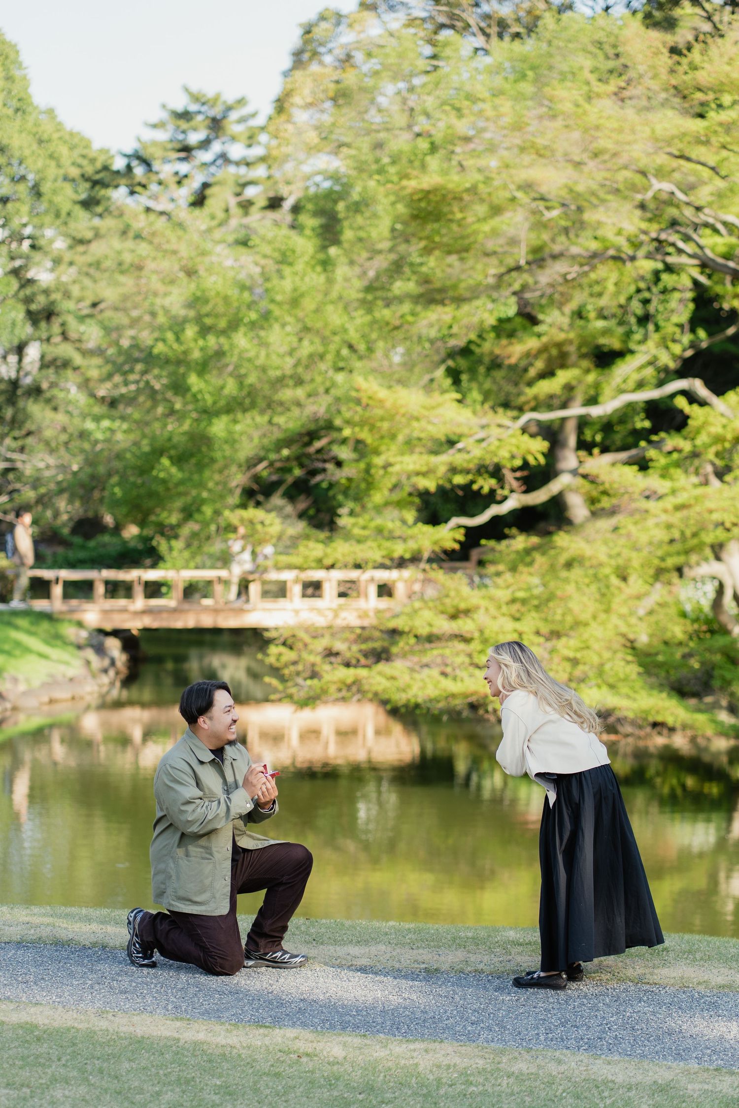Man proposes on one knee to woman in lush Japanese garden with pond and wooden bridge backdrop.