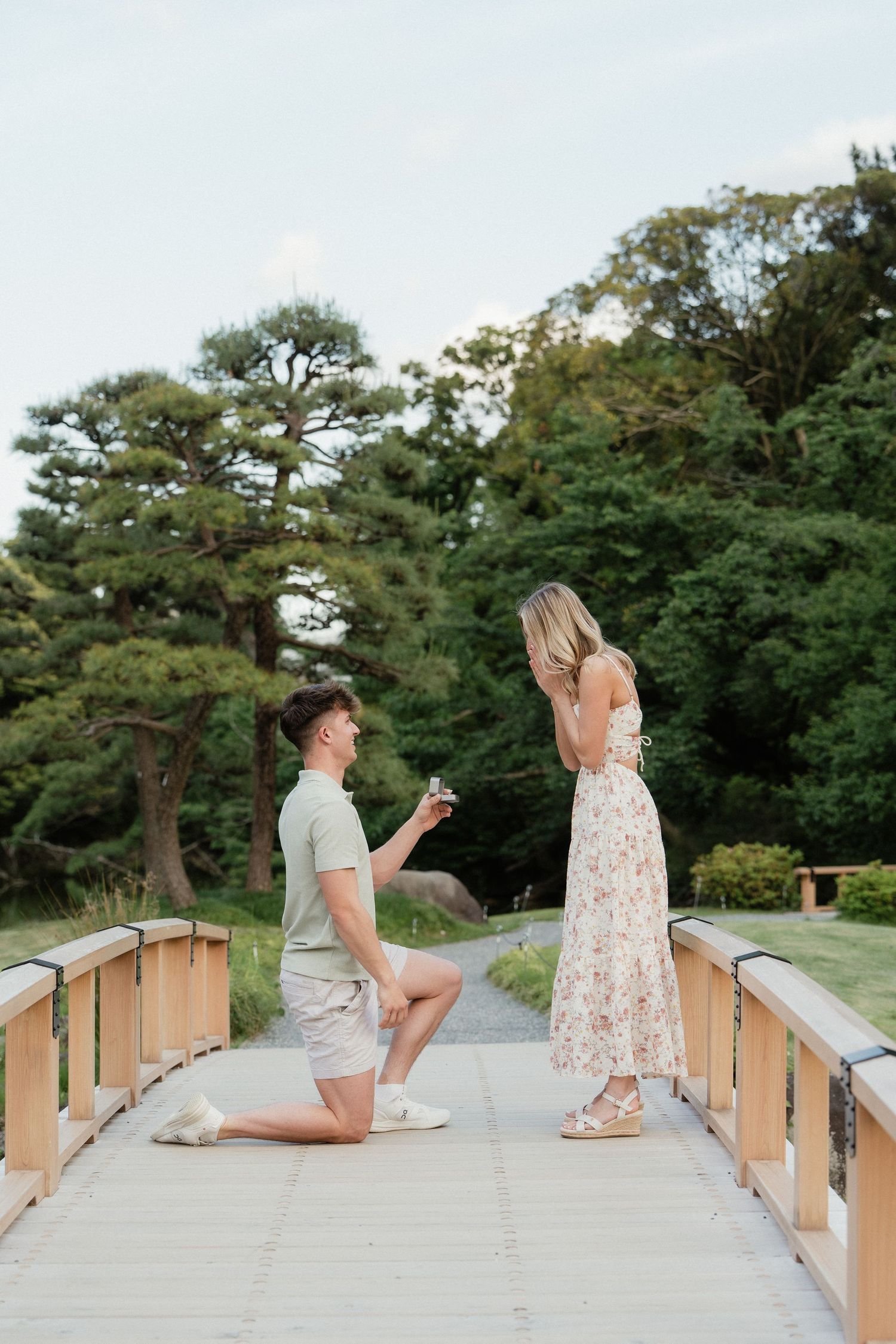 Man proposing on one knee to woman in floral dress on a wooden bridge surrounded by lush green trees.