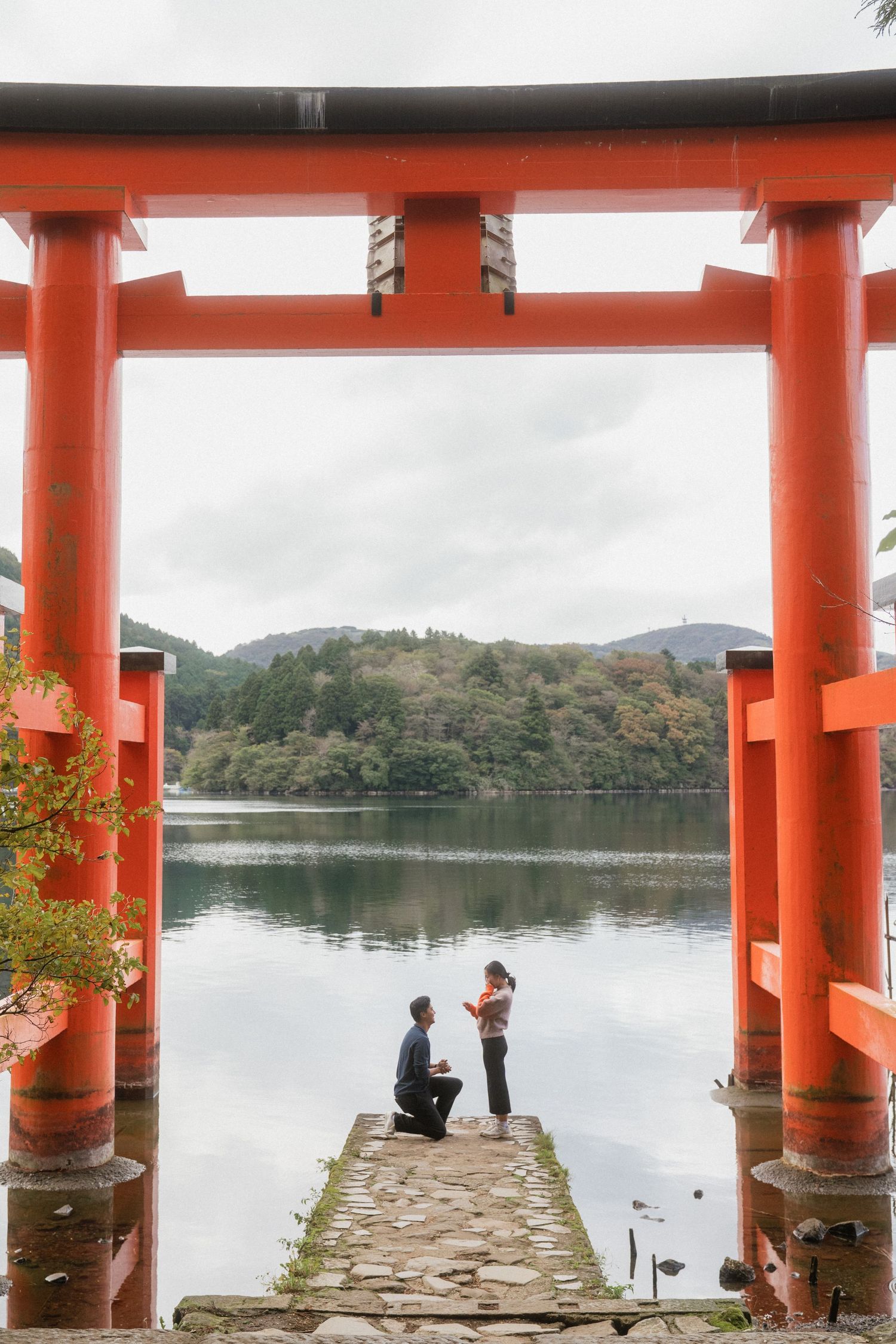 Man proposes to woman on stone pier framed by red torii gate at Lake Ashi, Hakone, Japan.