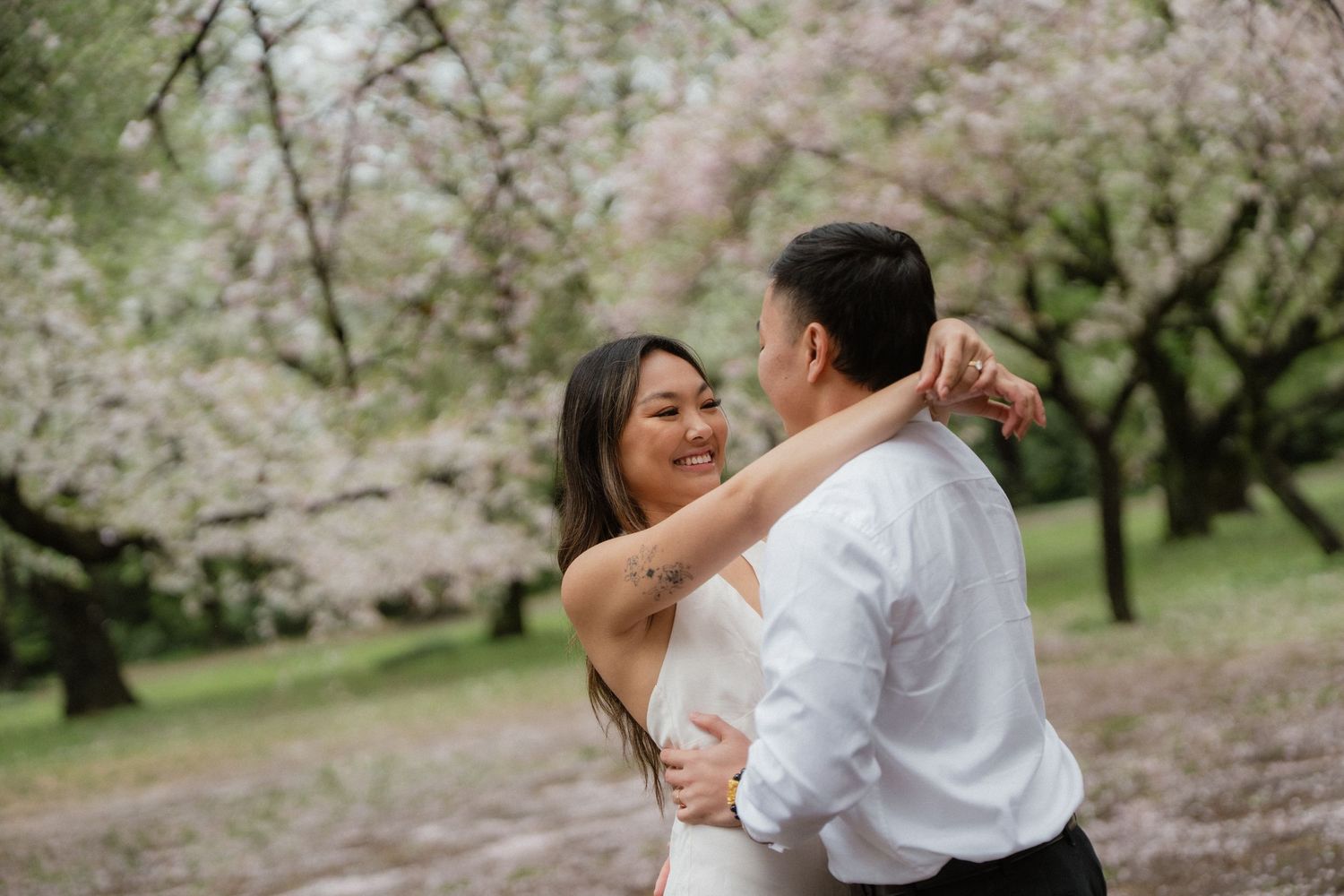 Couple dancing among blooming cherry blossom trees, woman in white dress smiling joyfully at partner outdoors.