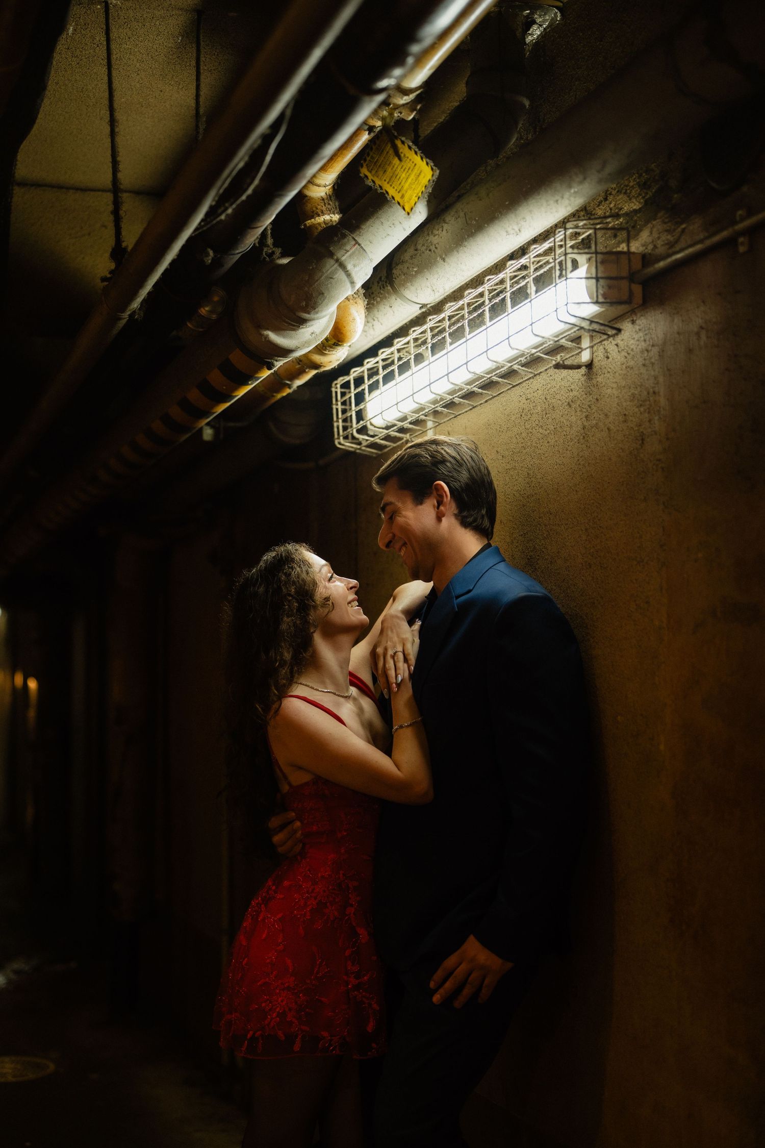 Couple in romantic embrace under industrial light in dimly lit corridor, woman in red dress, man in blue suit.