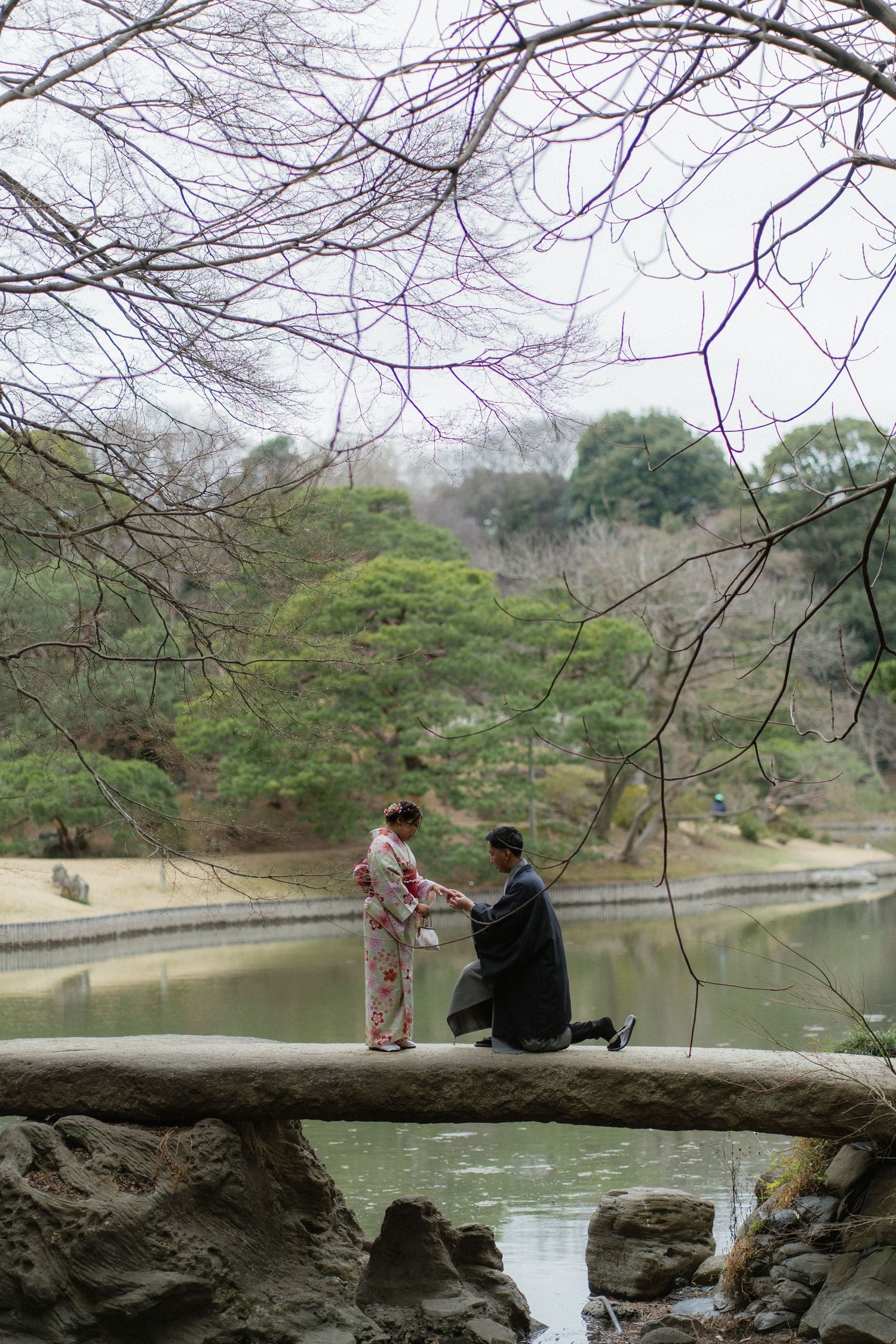 Man proposing to woman in kimono on stone bridge over pond in Japanese garden.
