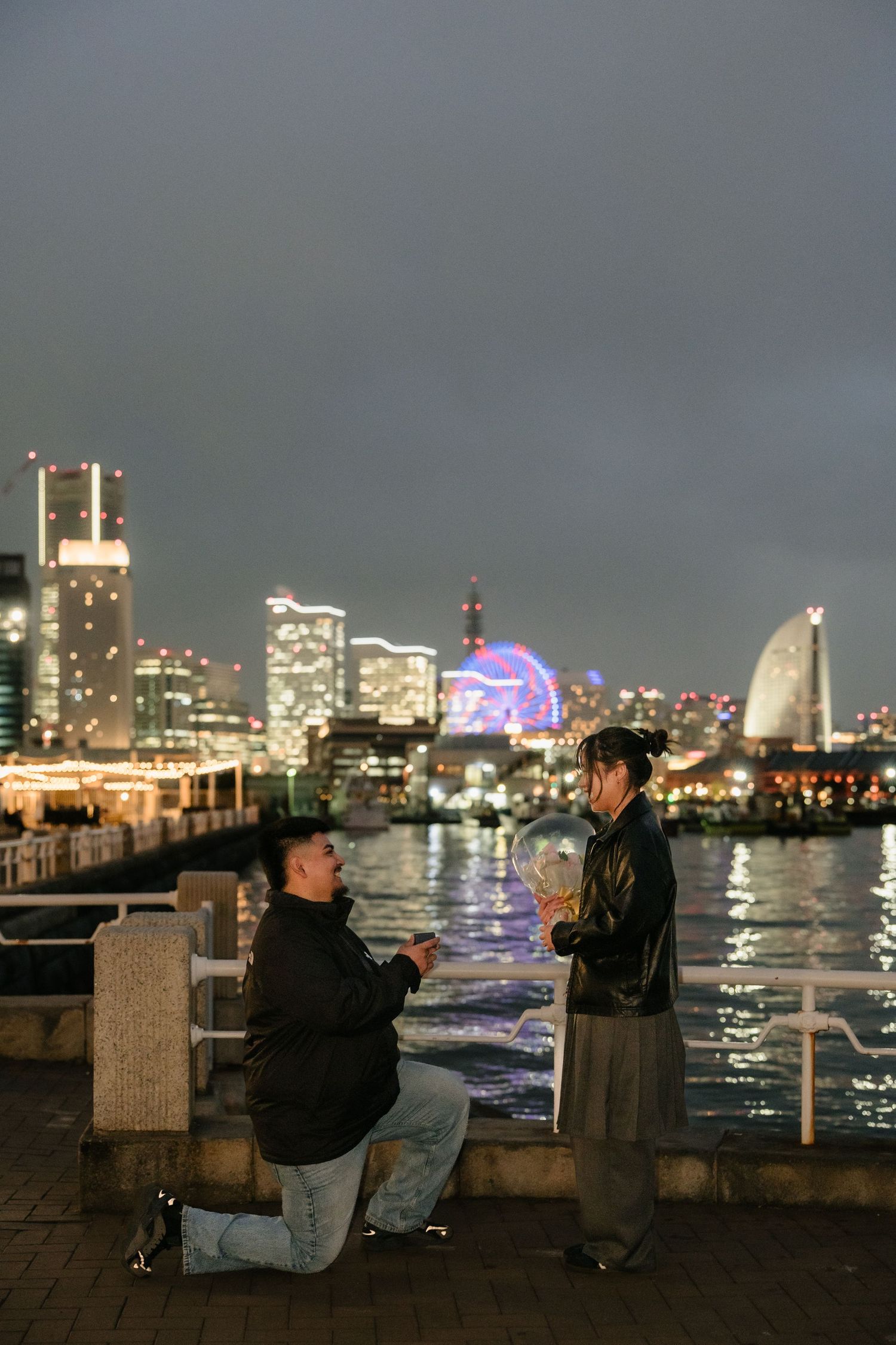 Man proposes on one knee to woman by waterfront with illuminated city skyline at night.