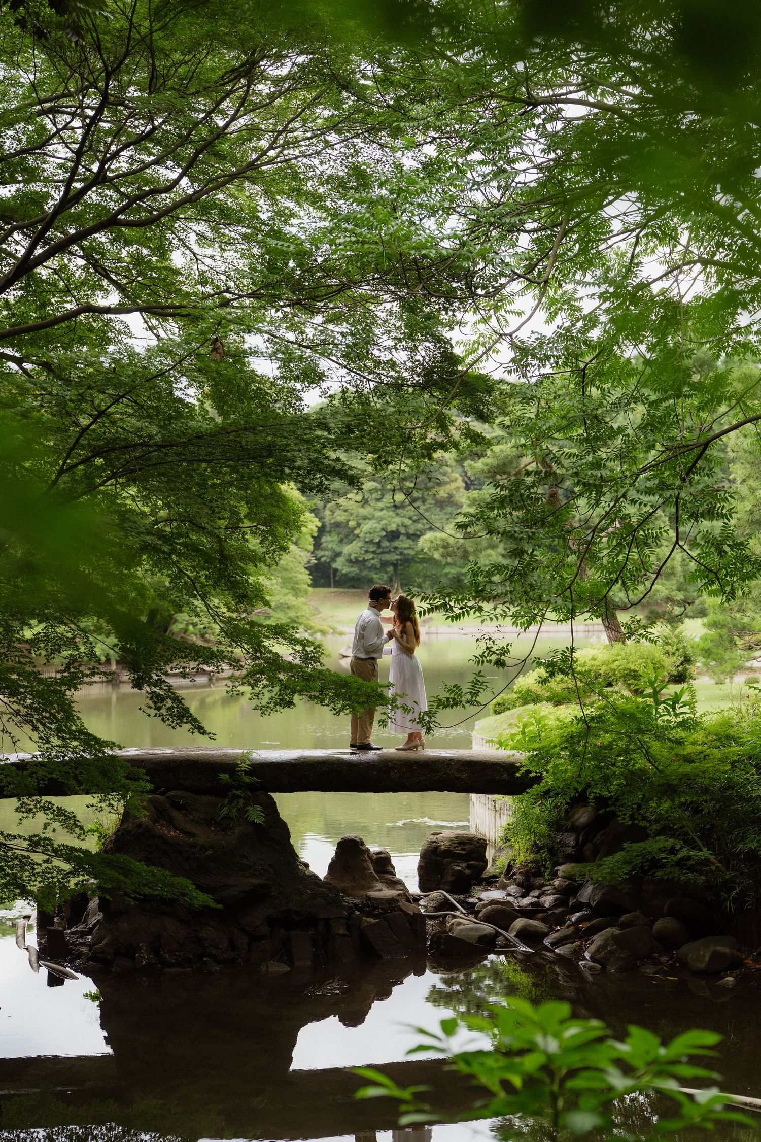 Couple standing on a stone bridge surrounded by lush green trees reflecting in a tranquil Japanese garden pond.