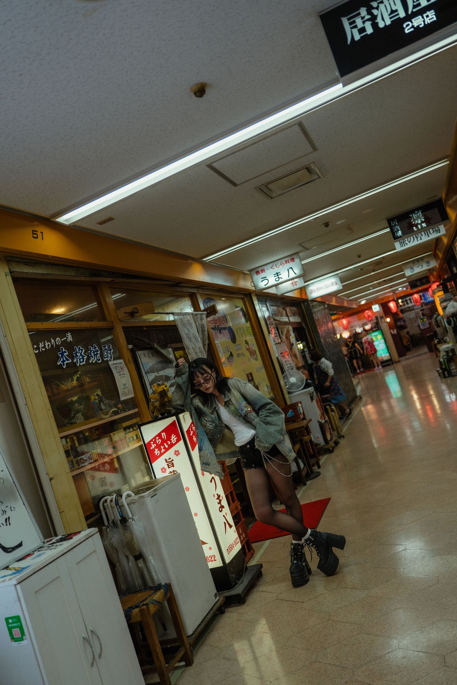 A young woman poses playfully in a Japanese indoor shopping arcade corridor with restaurant signs and storefronts.