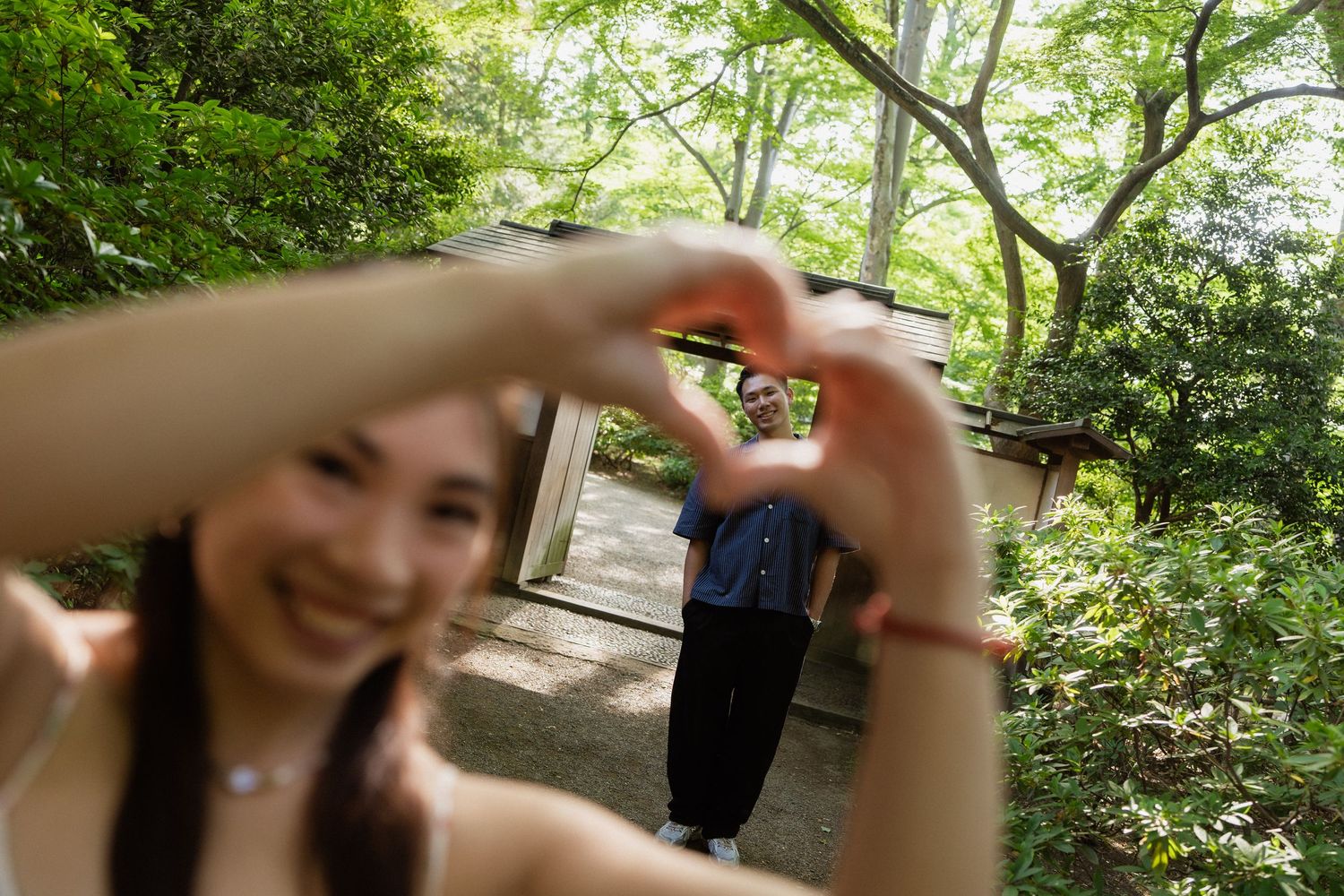 A smiling woman makes a heart shape with her hands while a person stands among lush green trees in the background.