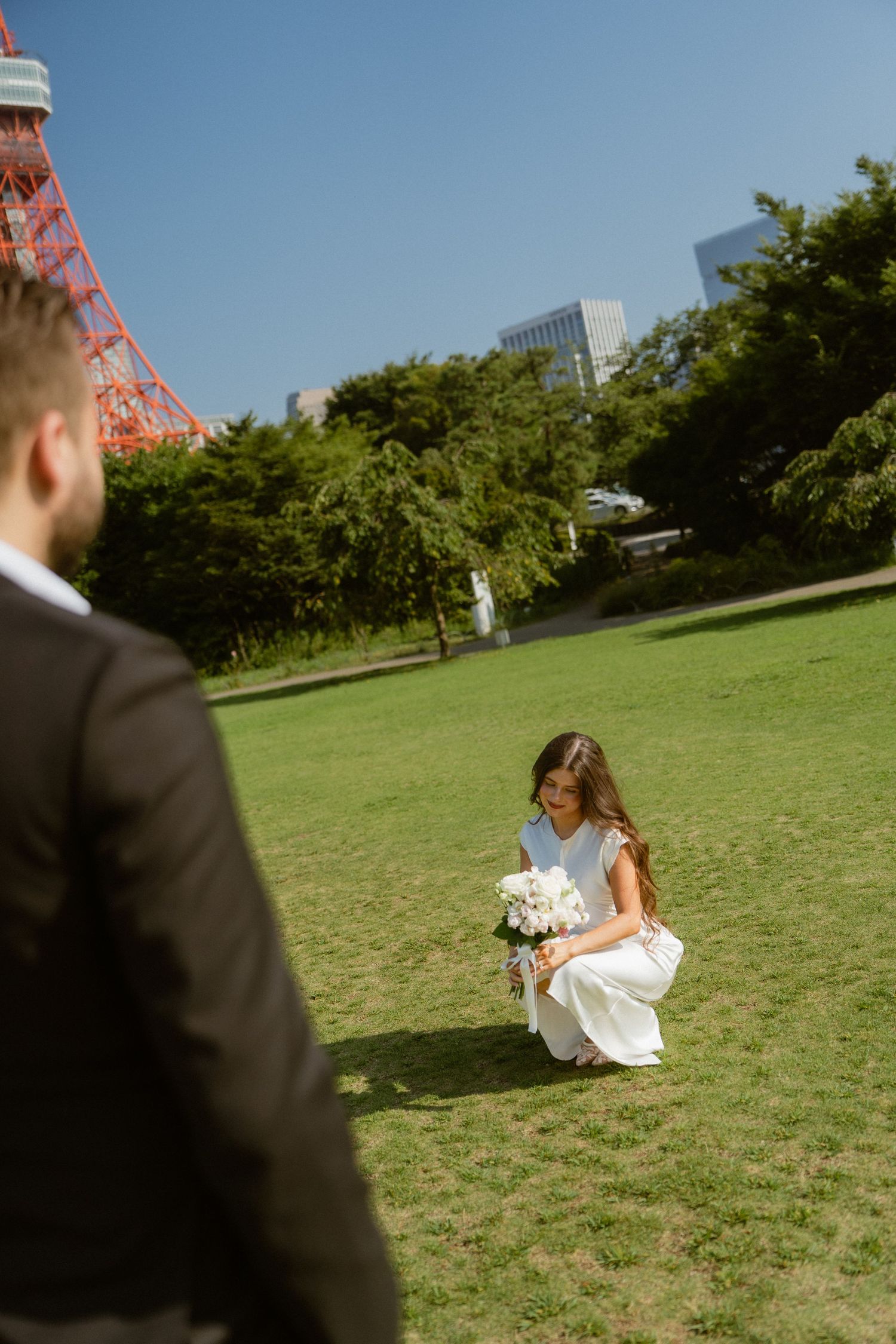 Bride in white dress crouching on green lawn holding bouquet near Tokyo Tower on sunny day.