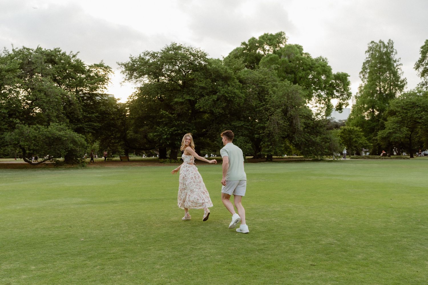 Couple dancing and holding hands on lush green lawn surrounded by tall trees at sunset.
